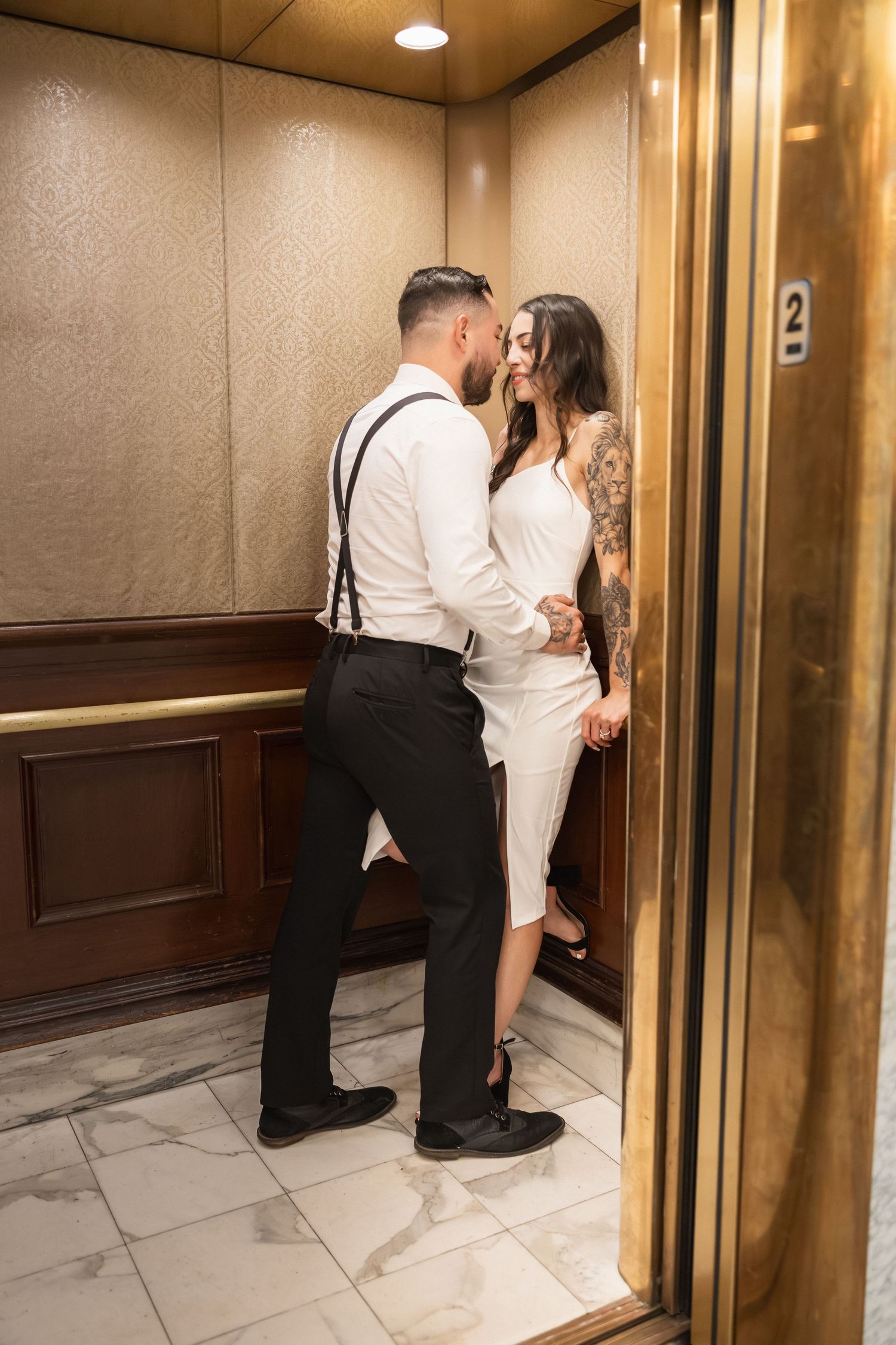 A romantic couple shares an intimate moment in an elevator, with the man wearing suspenders and woman in white dress.