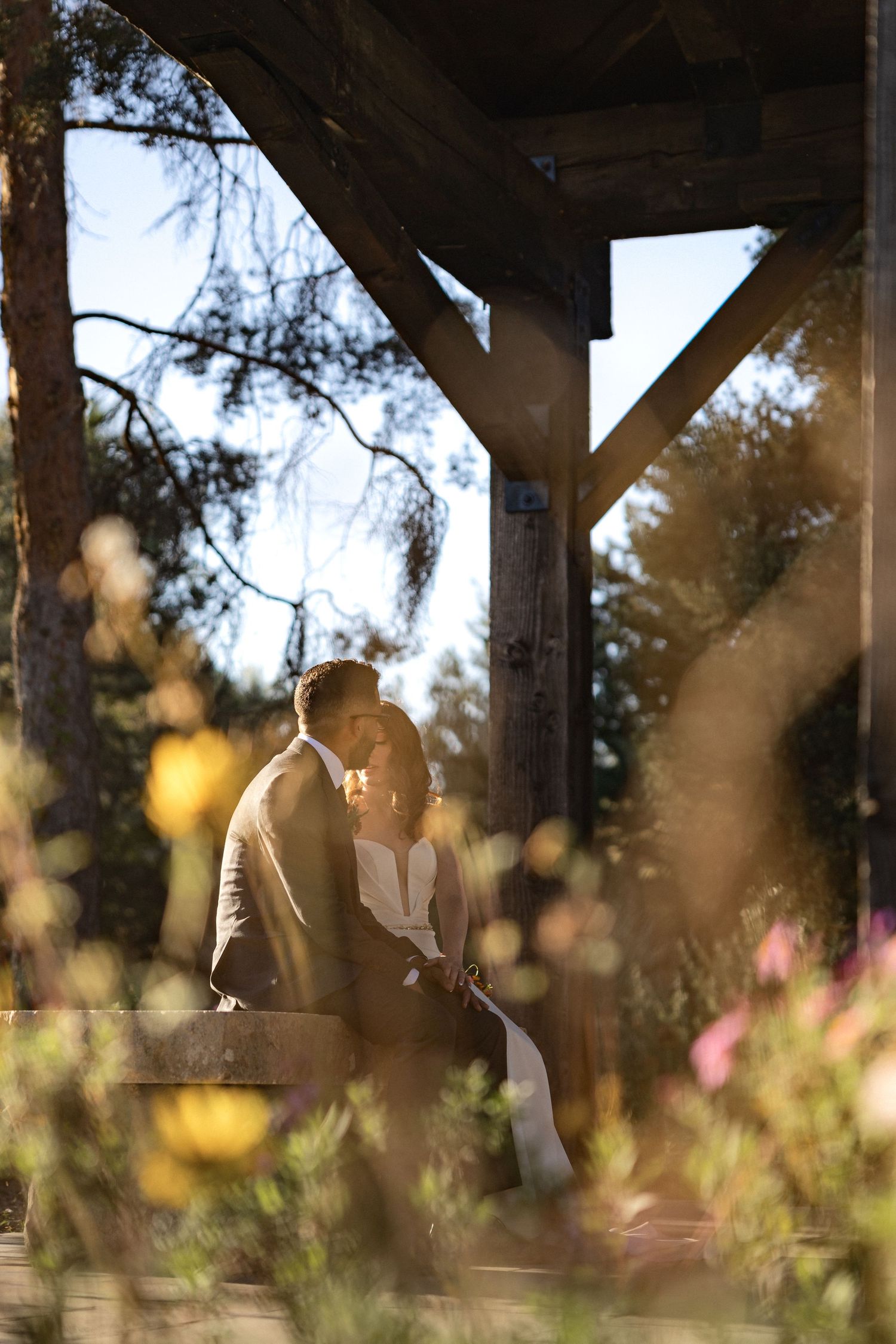 A couple in wedding attire sits on a stone bench in a garden setting during golden hour.