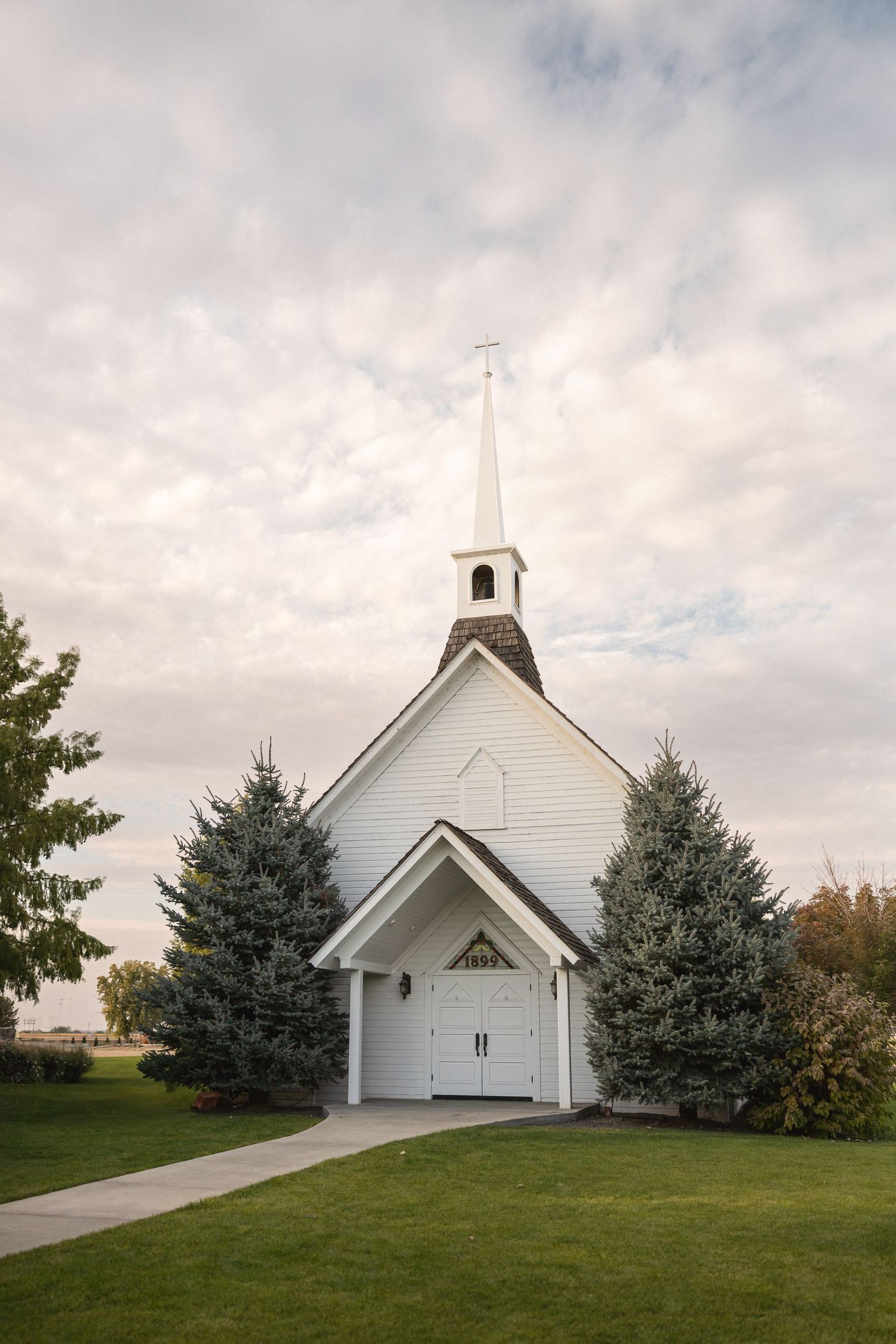 A pristine white chapel with tall spire surrounded by evergreen trees under cloudy skies.