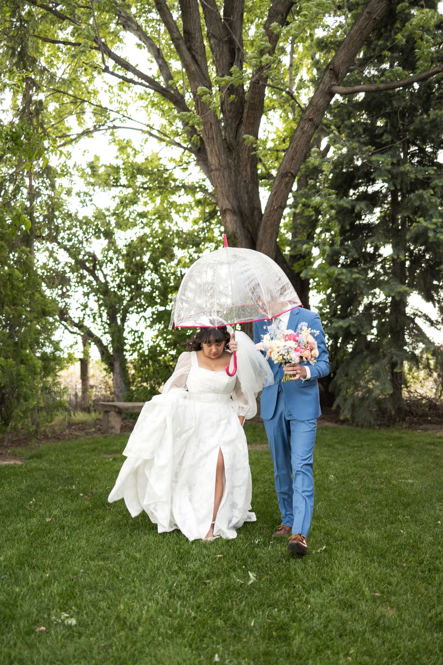 Wedding couple poses with clear umbrella in garden setting, bride in white dress and groom wearing bright blue suit.