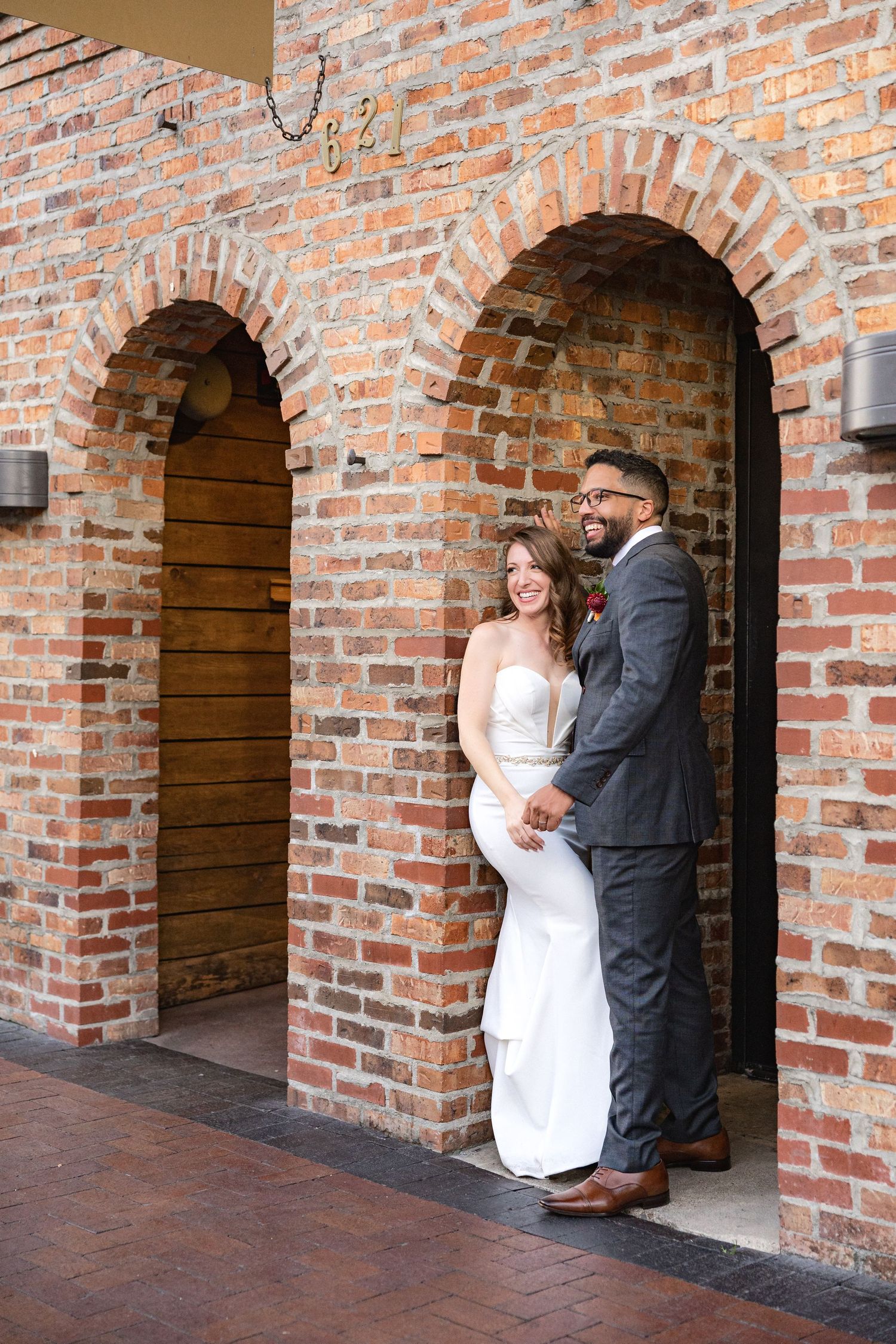 Newlyweds share a joyful moment while posing in a brick archway.