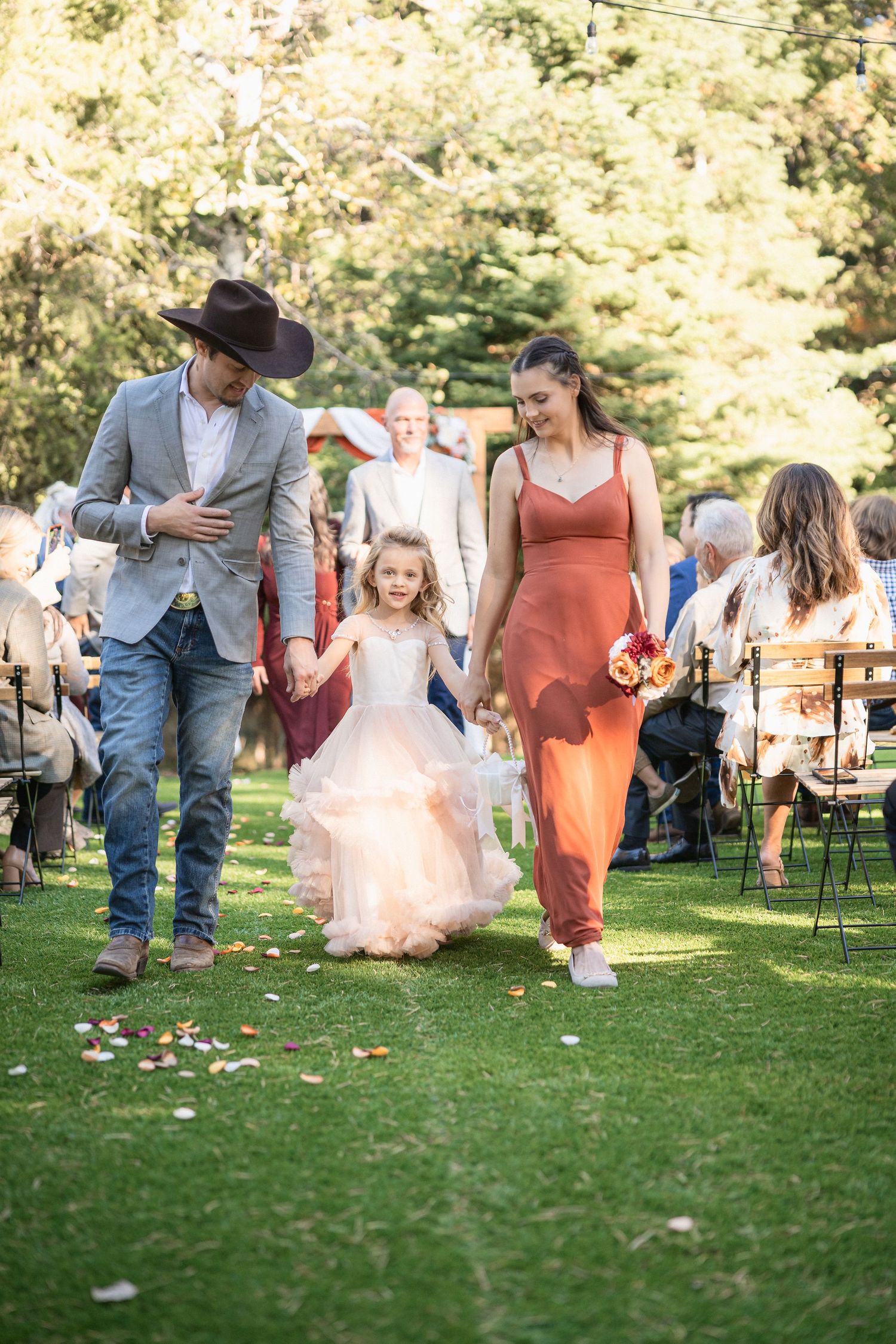 A couple in western wear walks with a young flower girl in a tulle dress during an outdoor celebration.