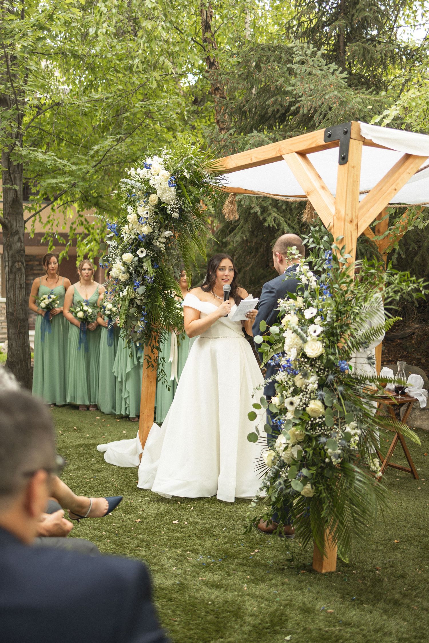 Wedding ceremony under wooden arch decorated with white flowers and greenery, with bridesmaids in sage dresses.