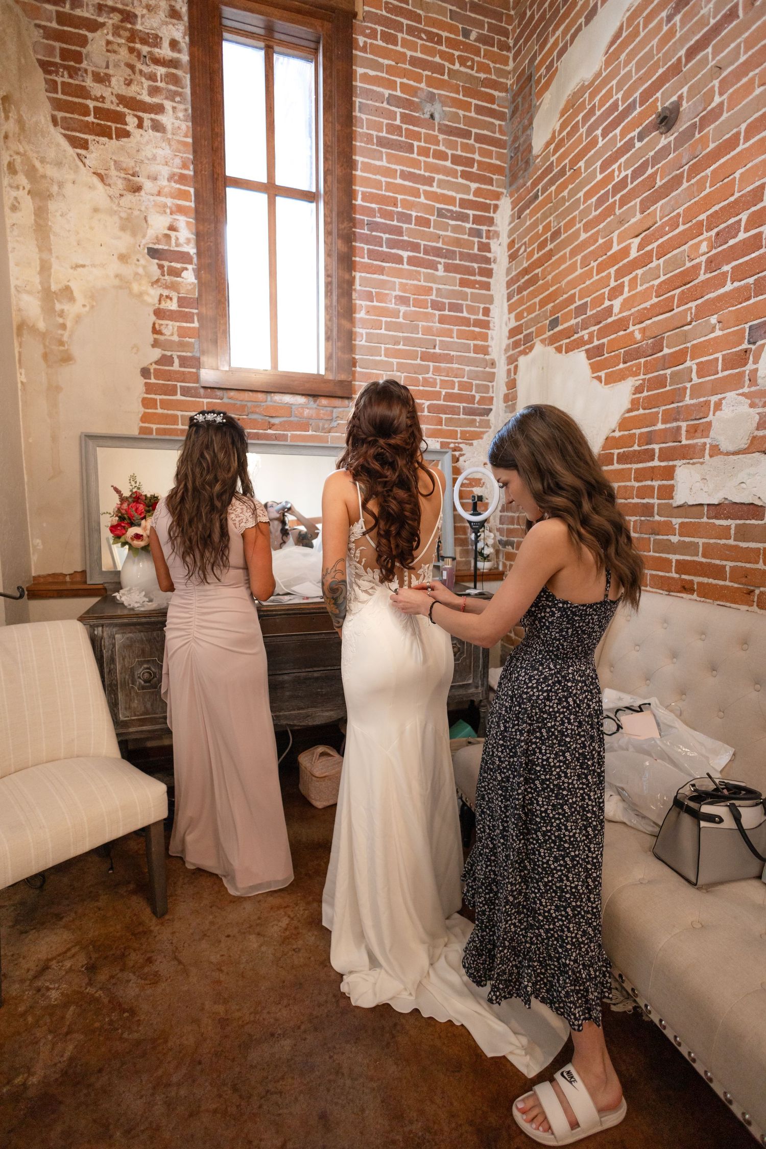 Wedding party helping with final dress adjustments in a rustic brick-walled room.