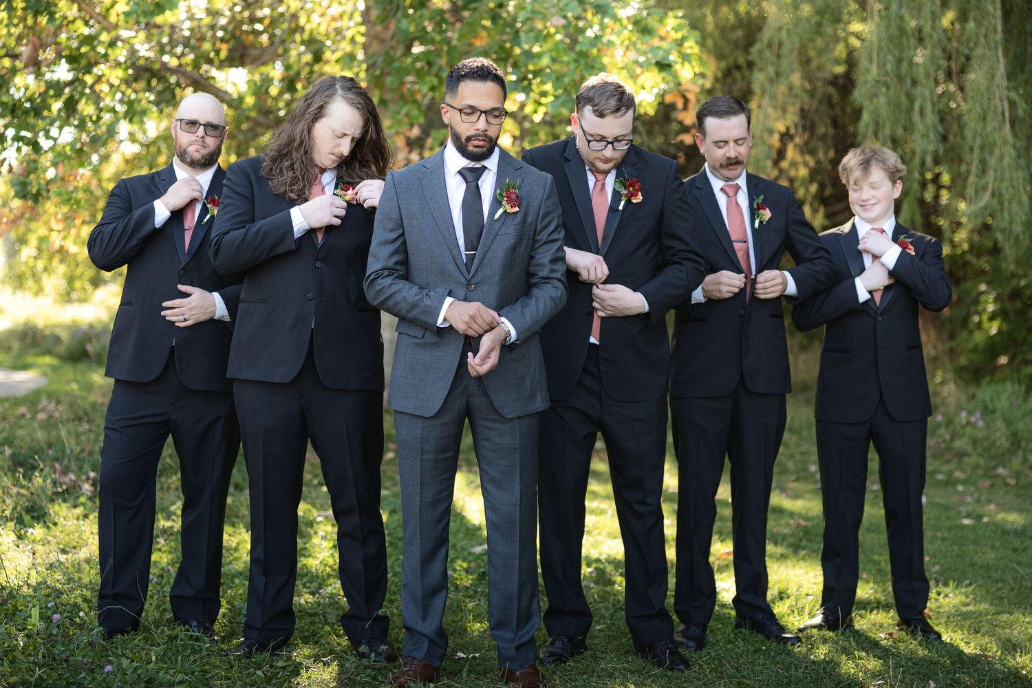 Wedding party groomsmen line up in formal suits against natural outdoor background.