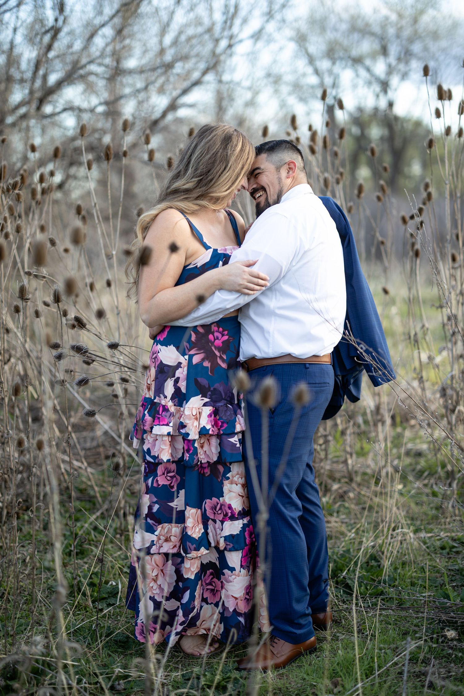 Couple in formal attire embraces among dried grass and cattails in a natural setting.