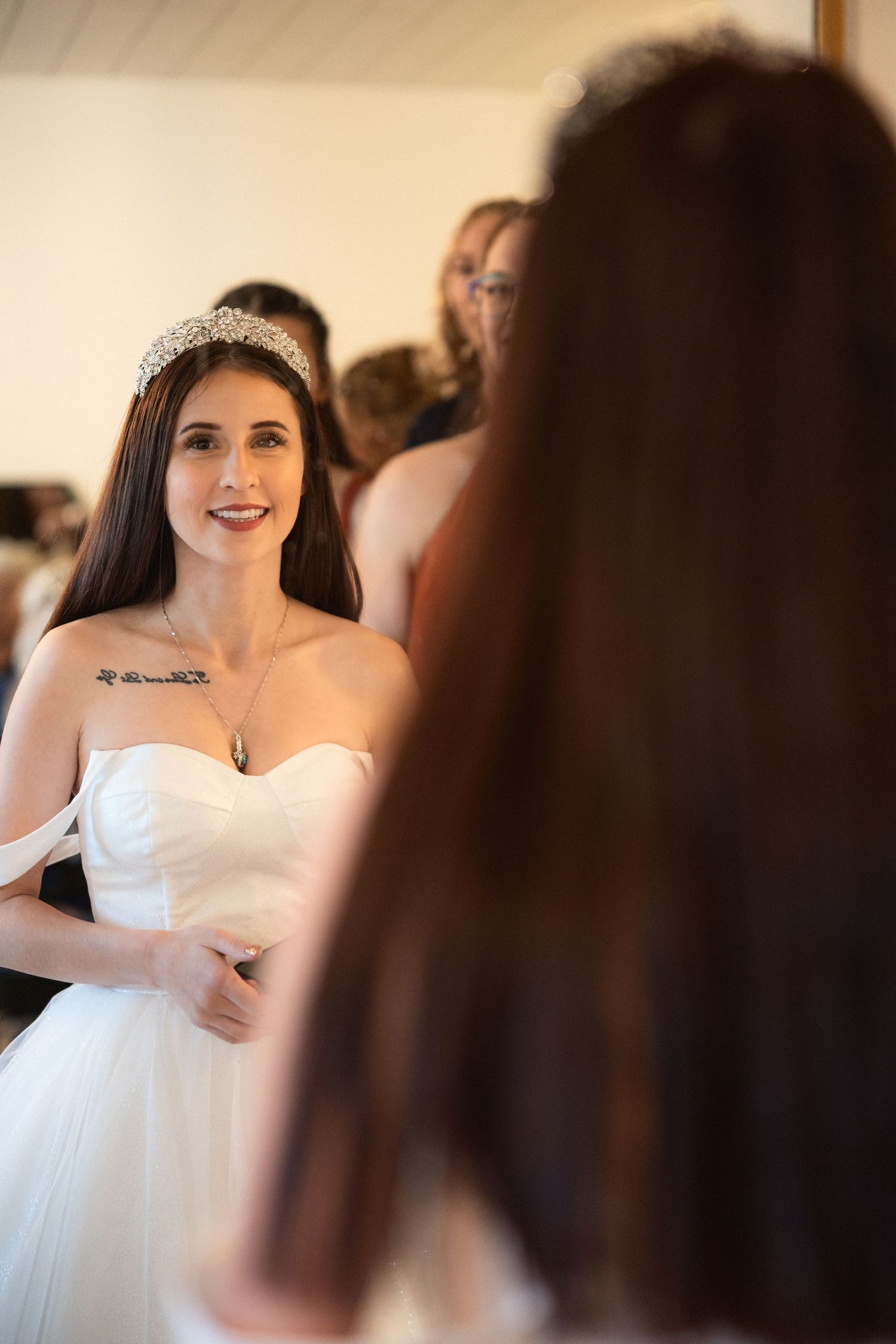 A bride wearing a white strapless gown and crystal tiara looks in the mirror during preparations.