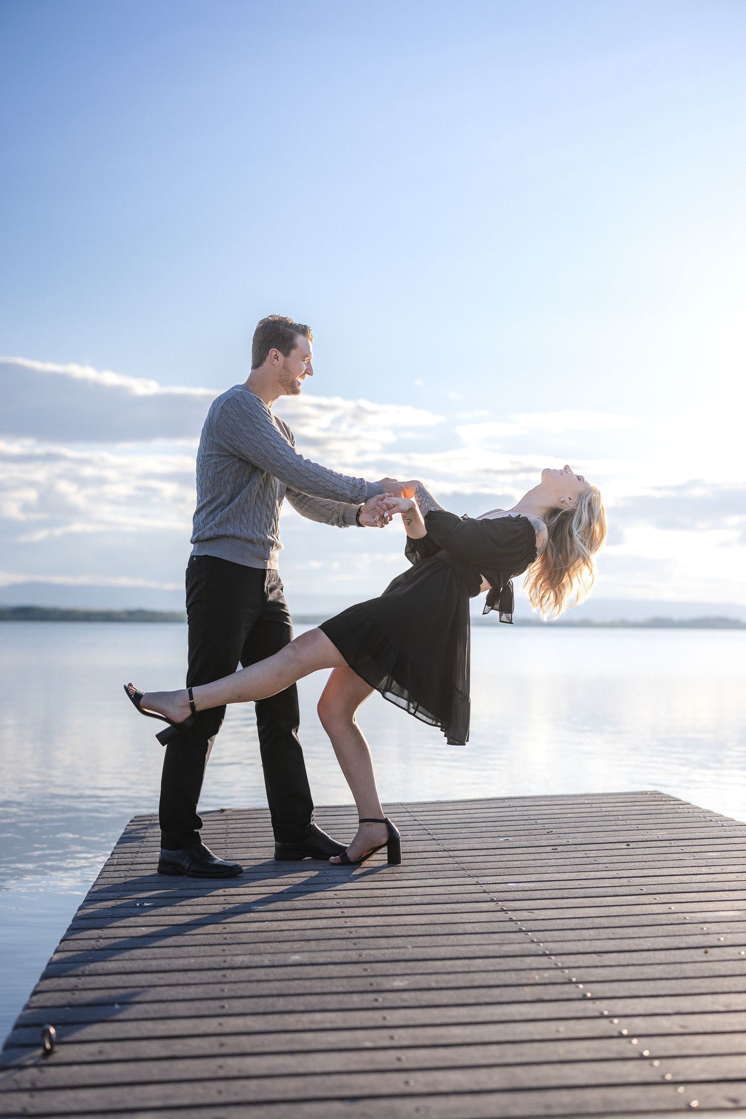 Couple performing dramatic dance dip on wooden pier with scenic lake background.