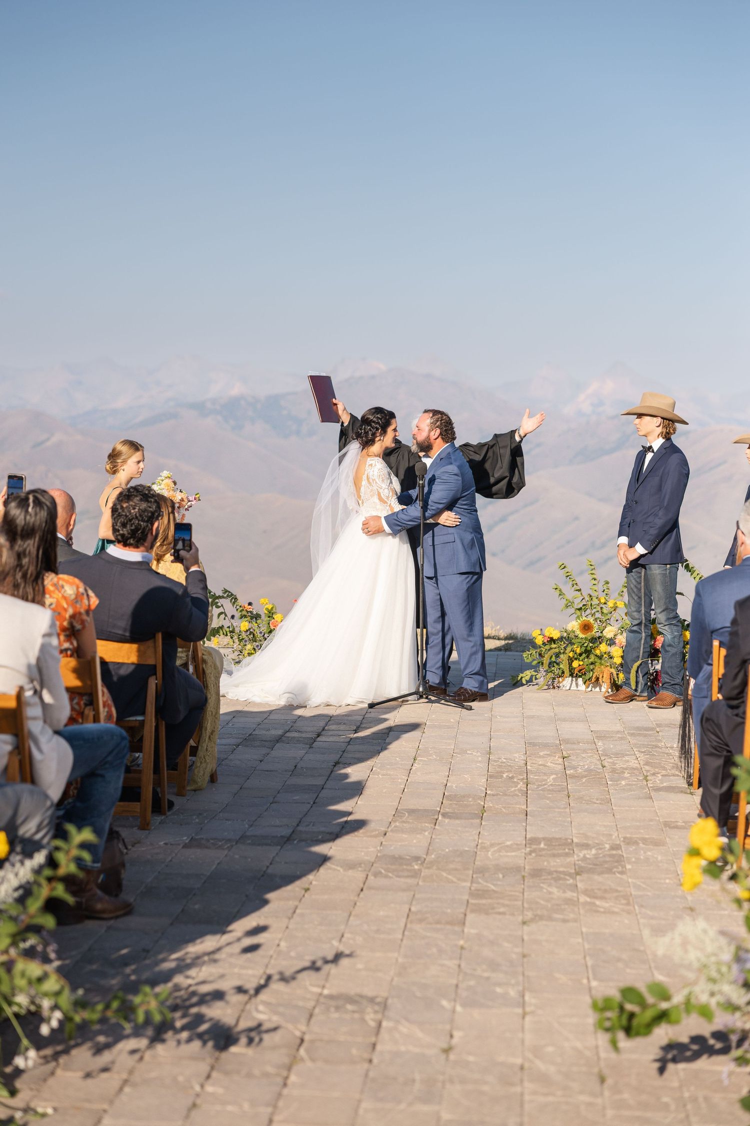 A wedding ceremony takes place on a stone patio with yellow floral decorations and mountain vistas.