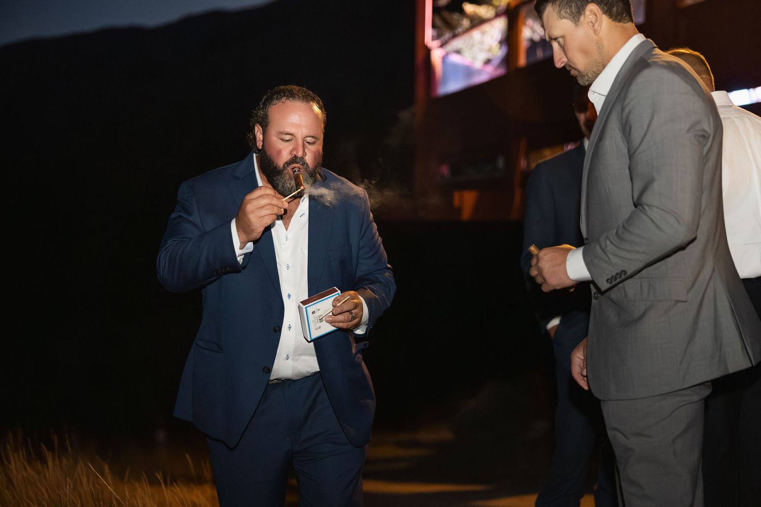 Wedding party members in formal attire sharing cigars outdoors at night.