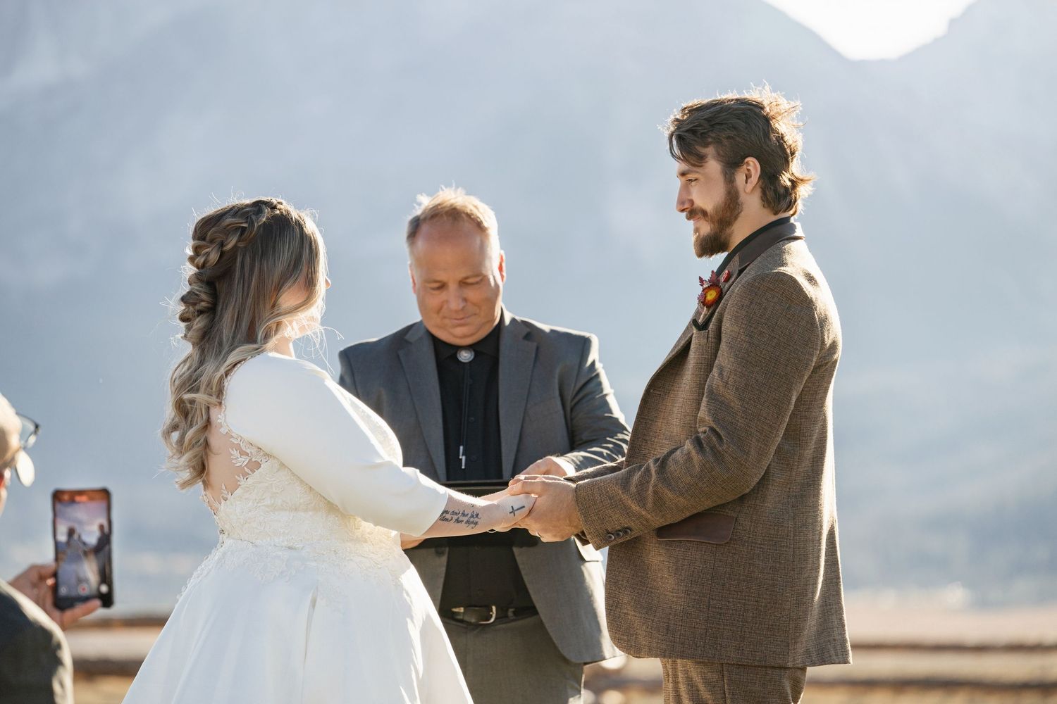 A scenic outdoor wedding ceremony with mountains in the background and guests seated on wooden benches.