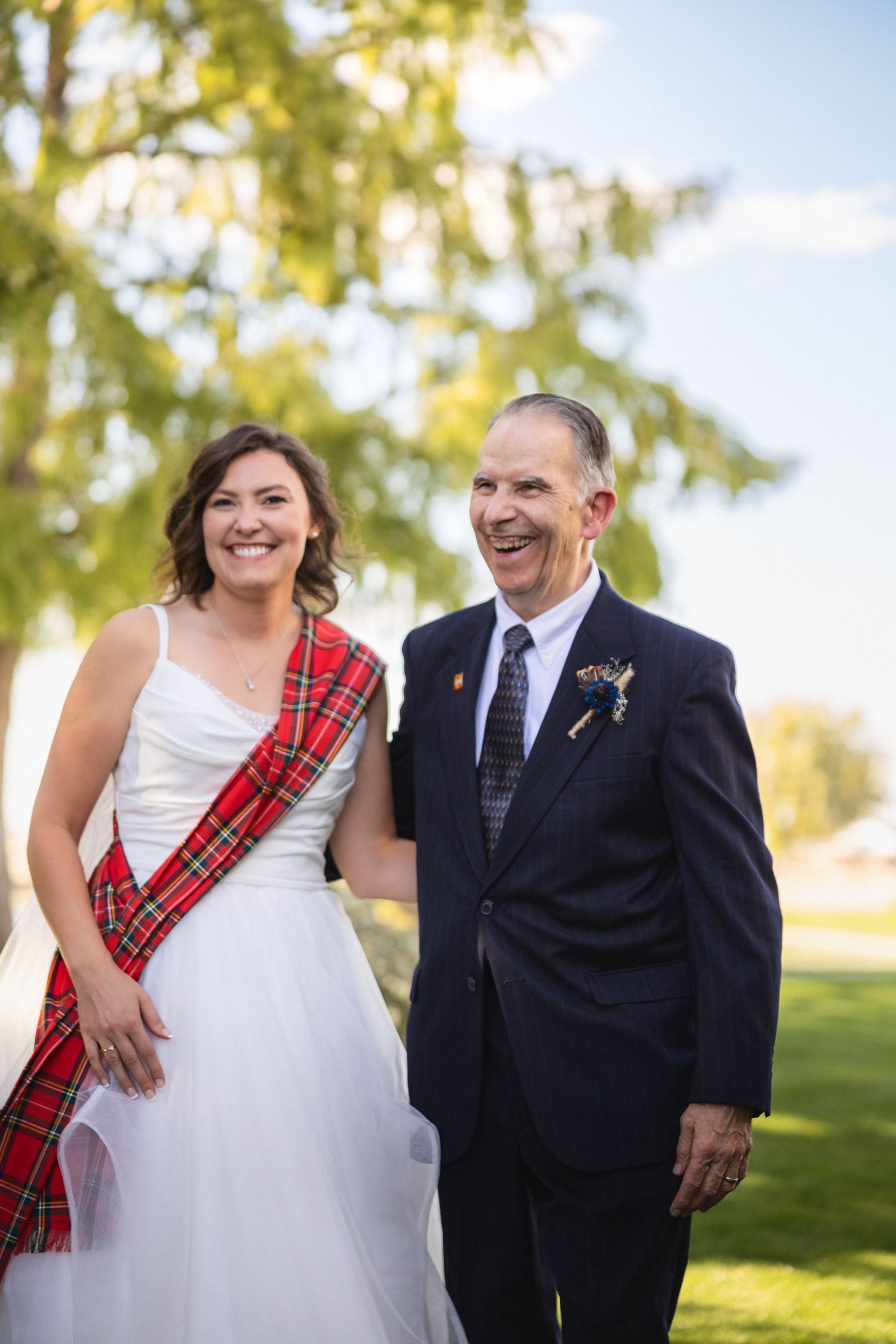 A joyful moment captured outdoors between two people, one in a white dress with tartan sash.