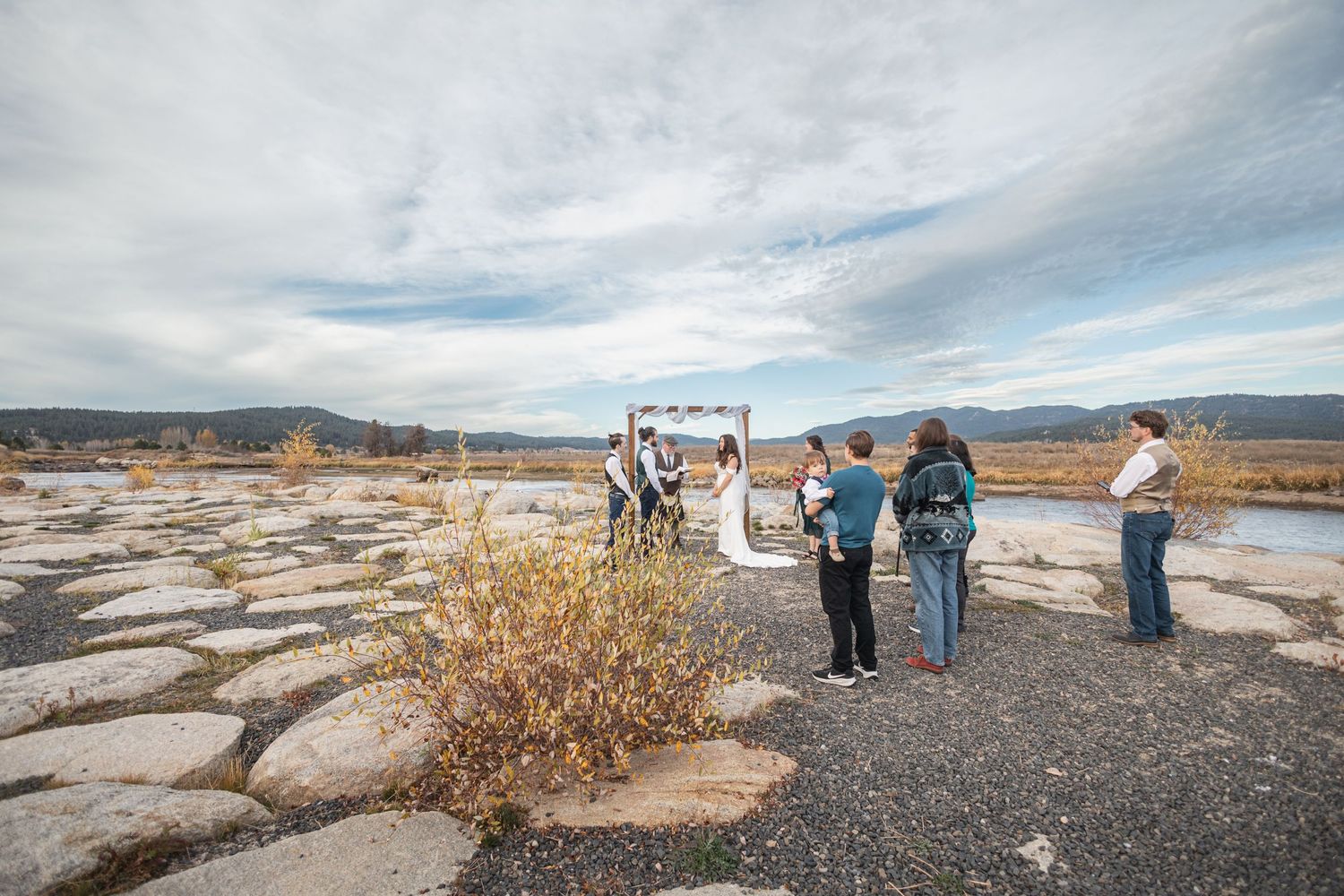 A wide-angle view of a small wedding ceremony on rocky terrain with mountains and cloudy skies in the distance.