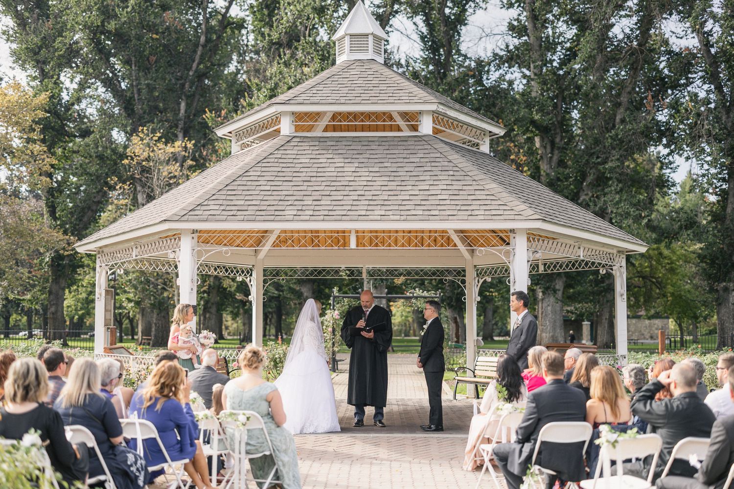 Wedding ceremony taking place in white gazebo with guests seated on white chairs.