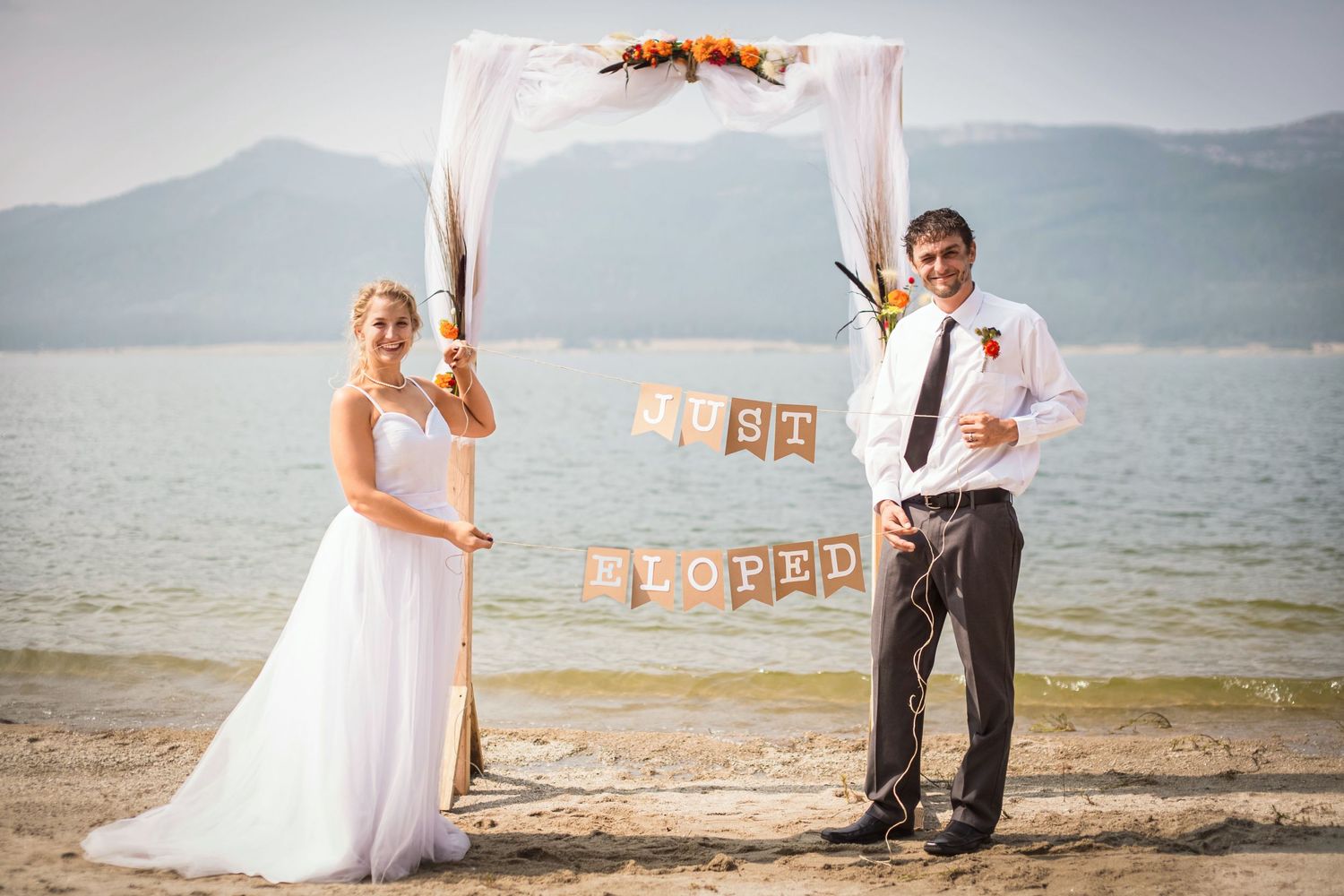Beach wedding couple posing with decorative signs under a floral arch by the water.