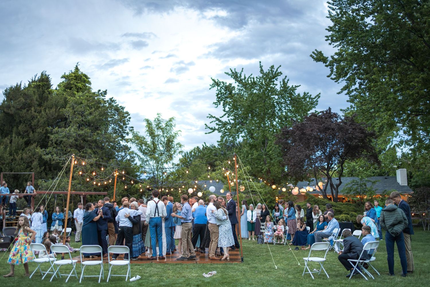 Guests gather at an outdoor wedding reception with string lights and white chairs.