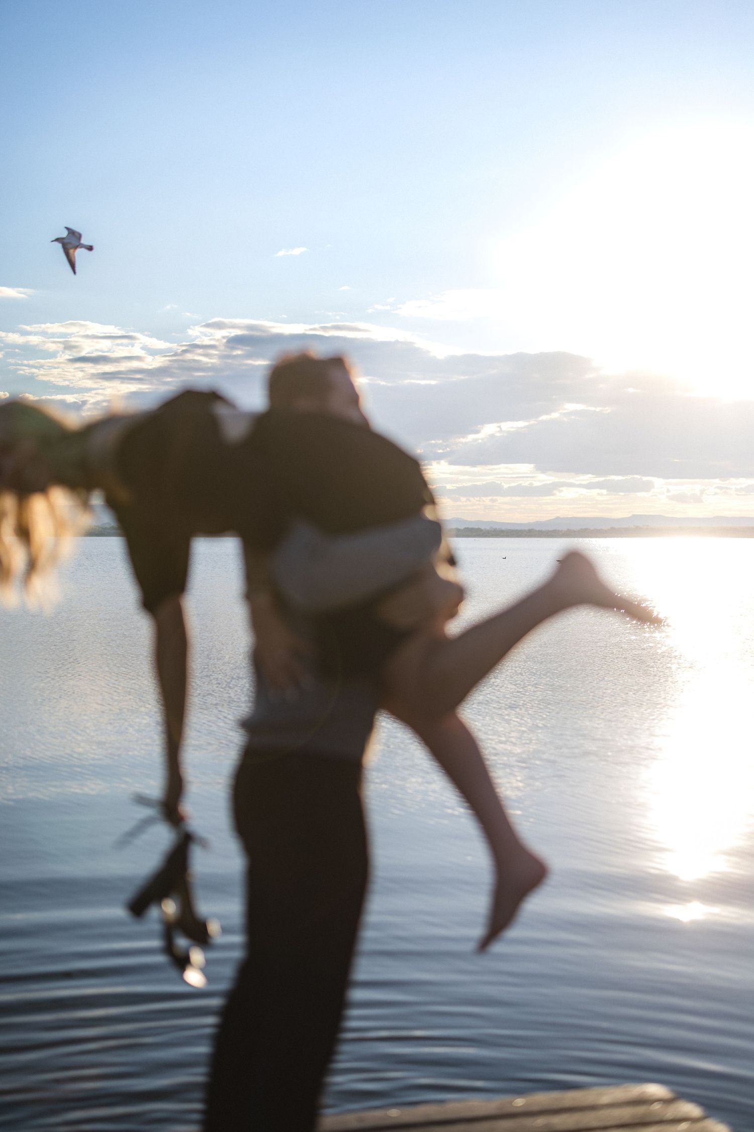 A couple performs romantic dance moves on a dock with water and sunset in the background.
