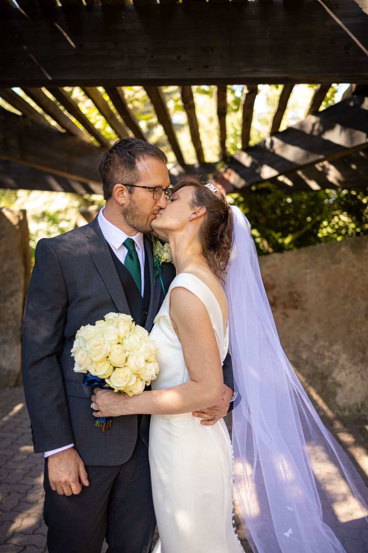 Romantic wedding portrait under wooden pergola with sunlit shadows.