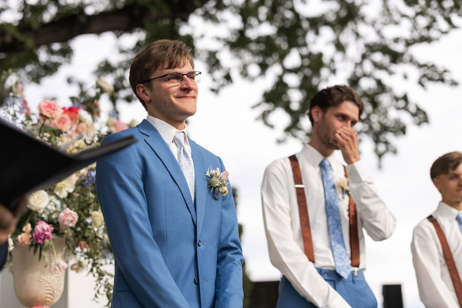 Groom in light blue suit and groomsmen in suspenders stand at outdoor wedding altar.