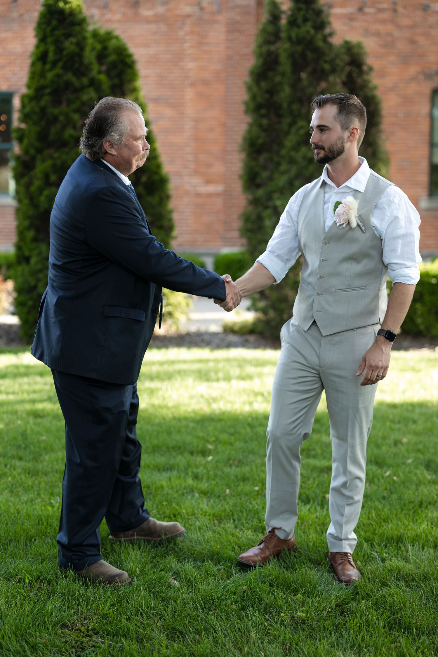 Groom in light gray vest shakes hands with man in navy suit on lawn.