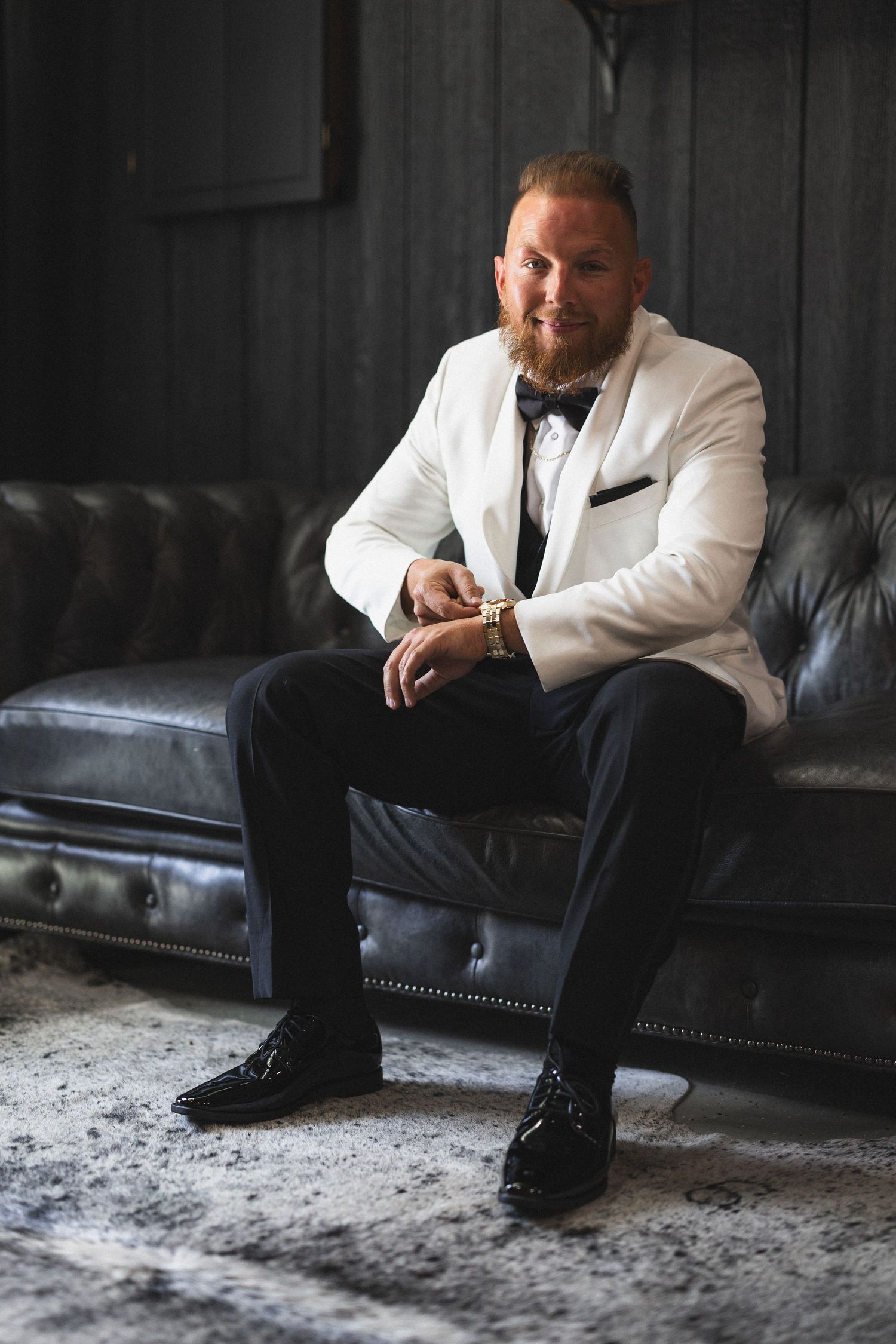 Groom in white tuxedo jacket sits confidently on leather couch before wedding.