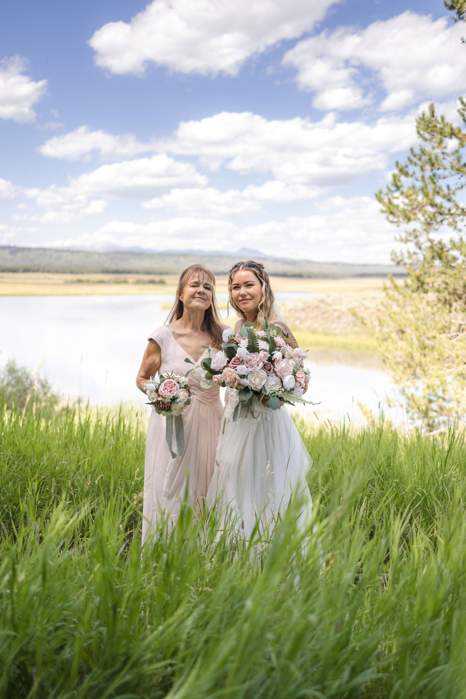 Wedding party members in light dresses holding floral bouquets stand together in a meadow with mountains in the background.