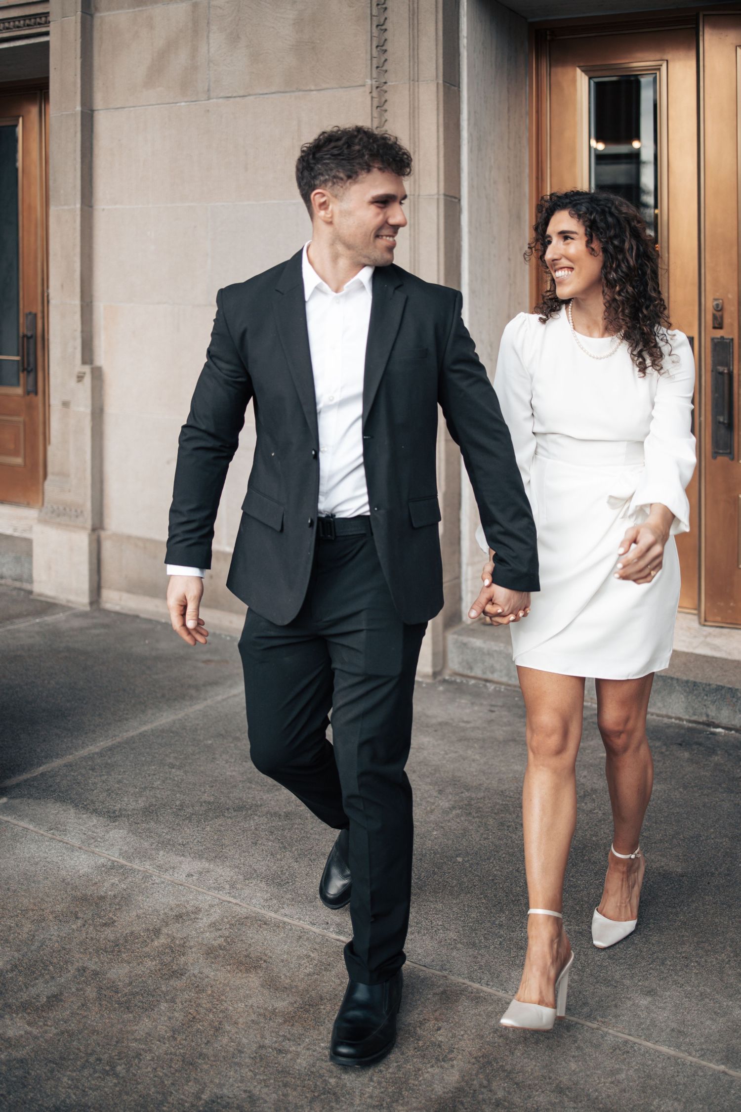 Stylish couple in formal wear walks together on urban sidewalk outside historic building.