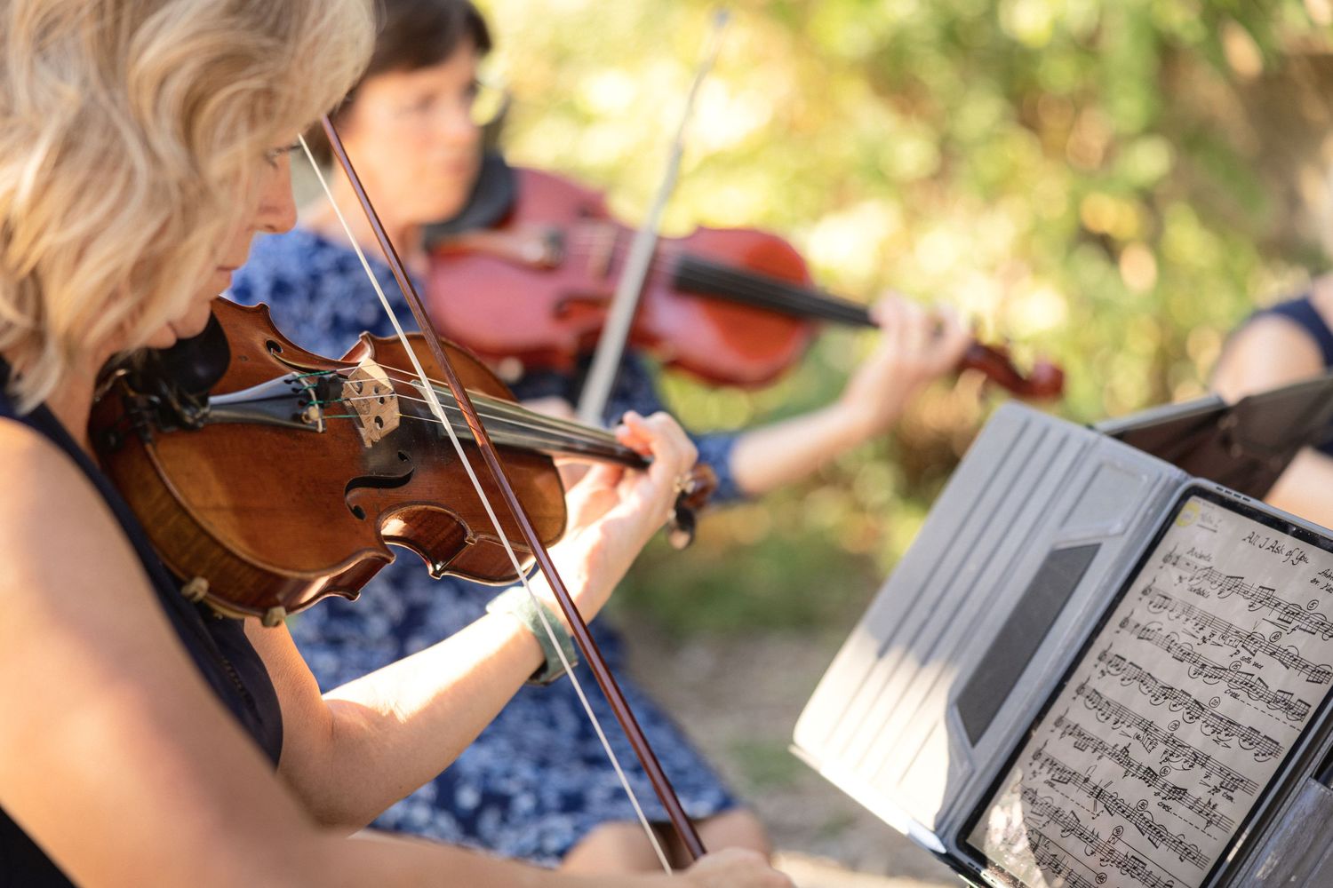 Musicians perform on violins outdoors with sheet music on stands.
