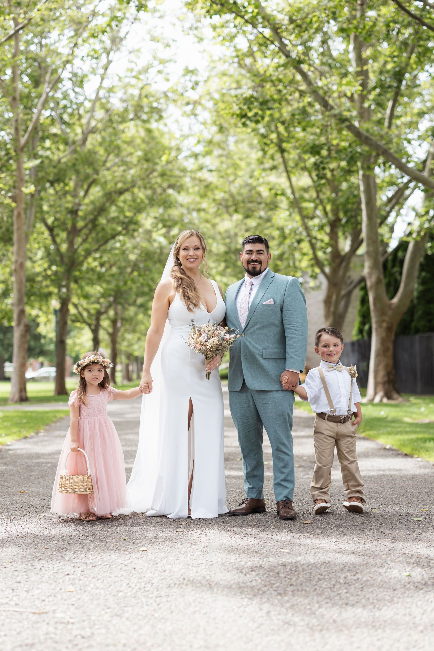 Wedding party with bride in white gown, groom in blue suit, and two children pose on outdoor path.