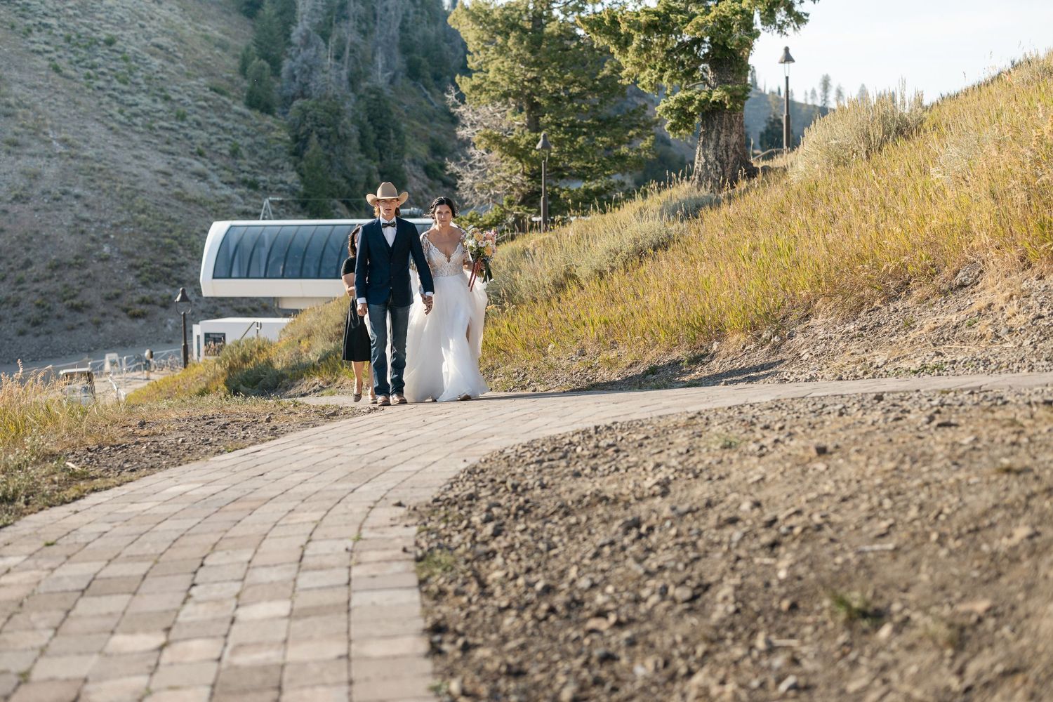 Couple walks down a stone pathway with mountains and ski lift visible in background.