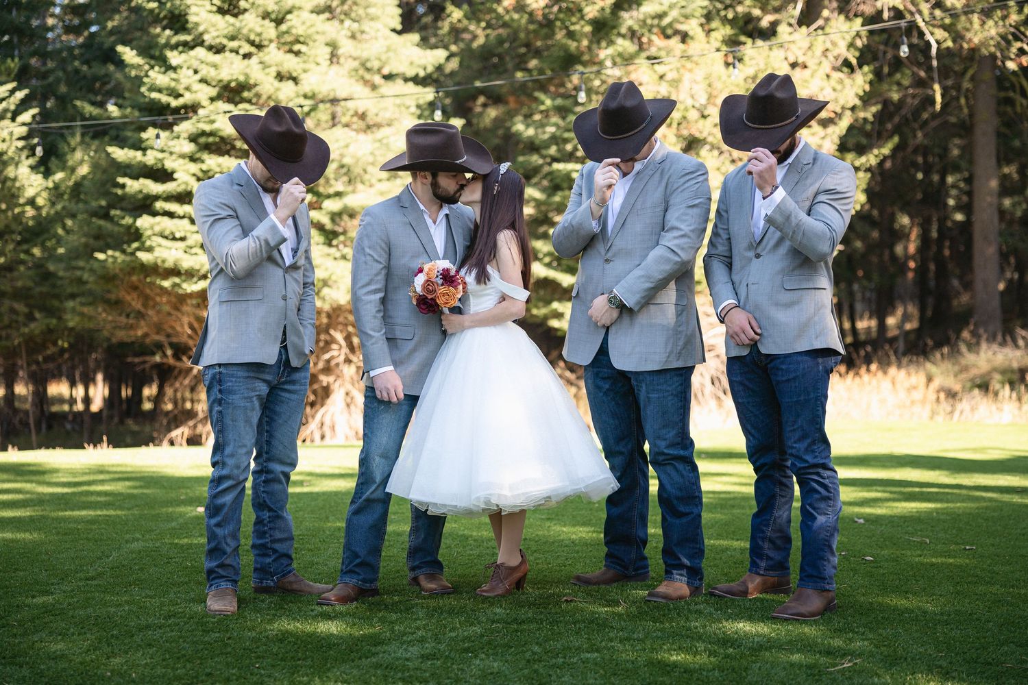 Groomsmen in cowboy hats and light blue jackets pose with bride in short wedding dress on grassy lawn.