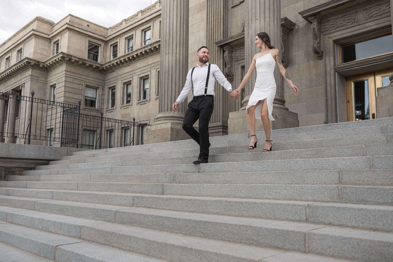 A couple poses playfully on grand stone steps of a neoclassical building with columns.