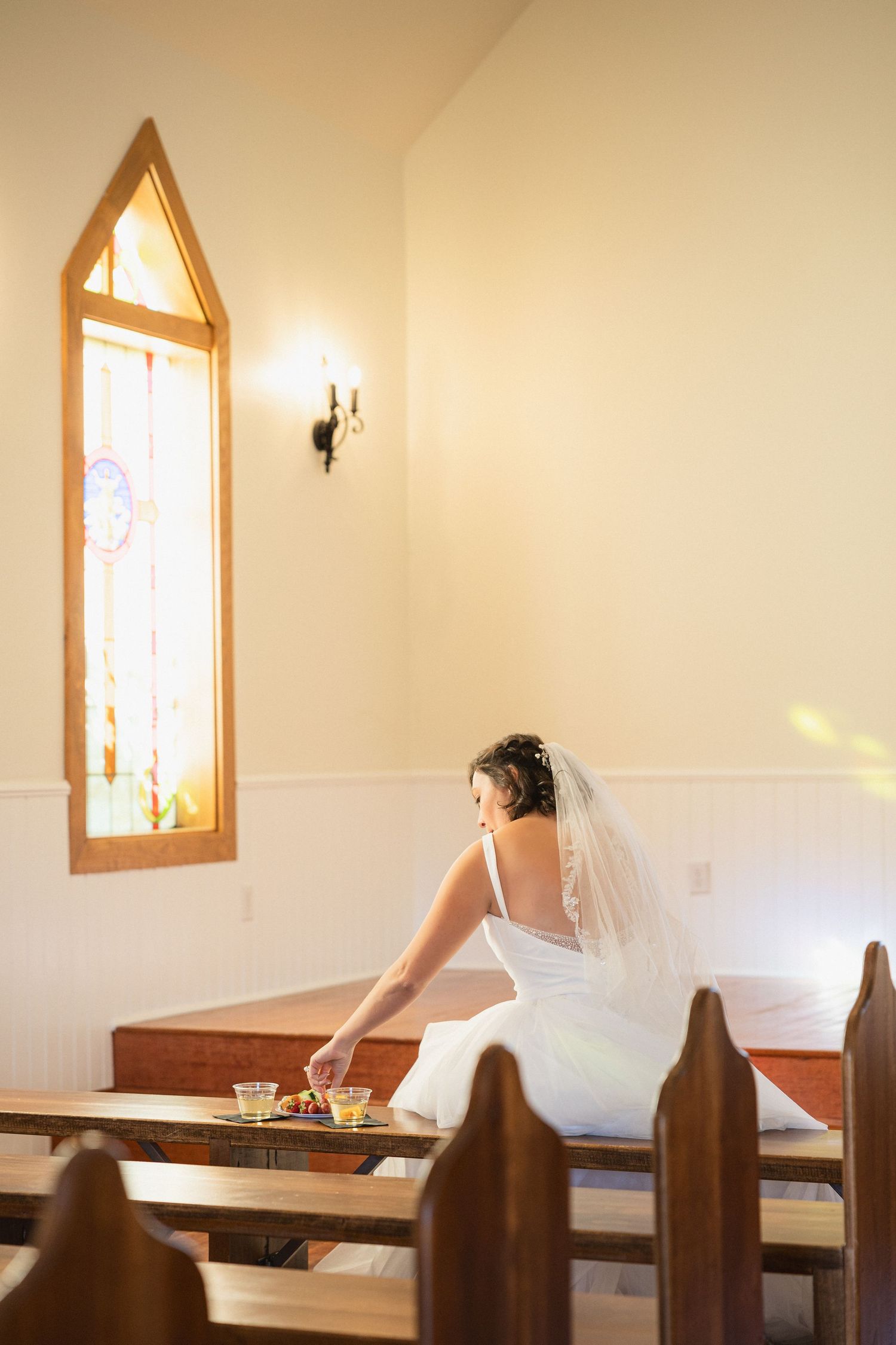 Bride in white dress sitting alone in a chapel pew near a stained glass window.