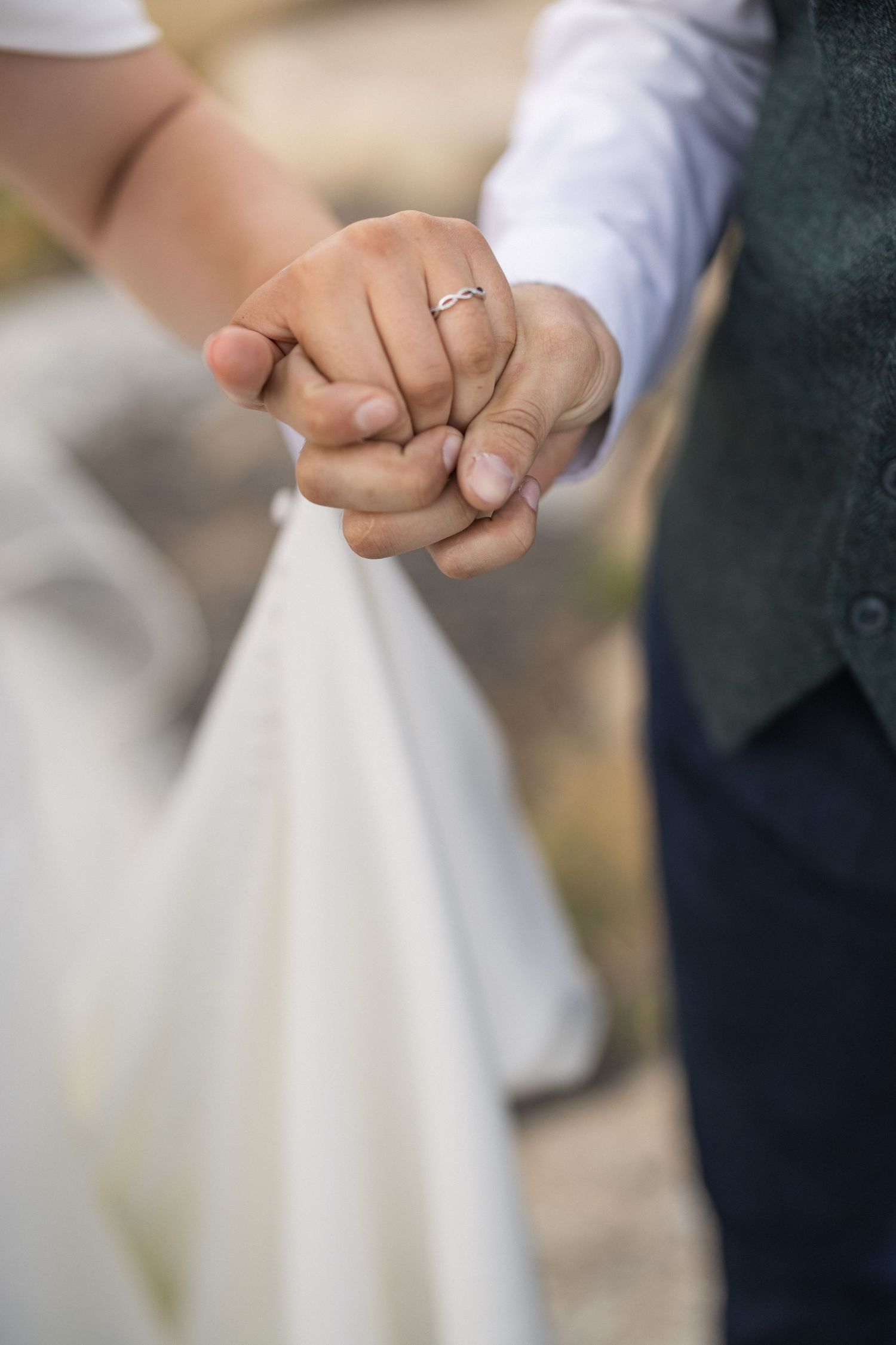 Close-up of joined hands with engagement ring against white dress and dark vest.