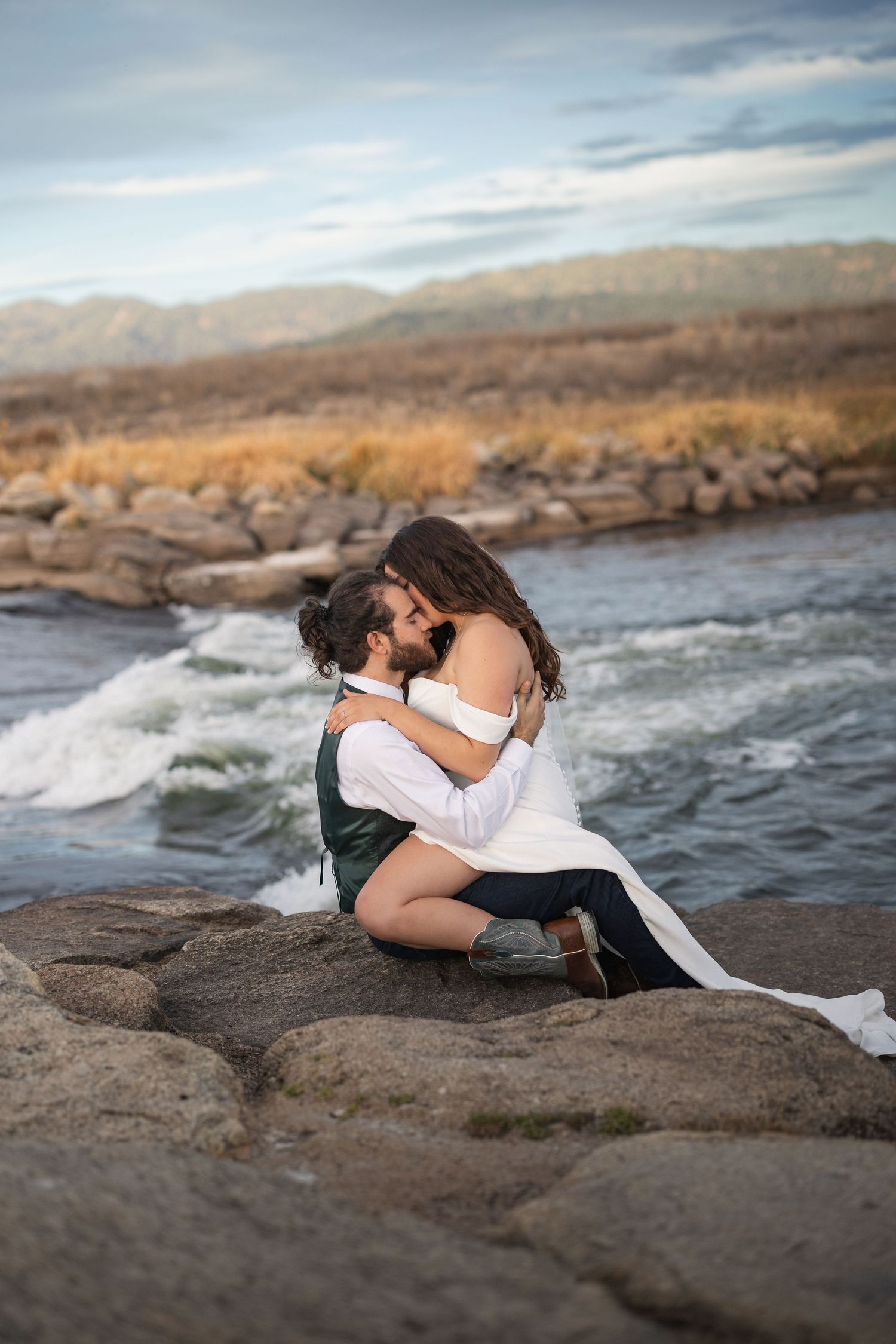 Couple in wedding attire embracing on riverside rocks with flowing water and mountains behind.