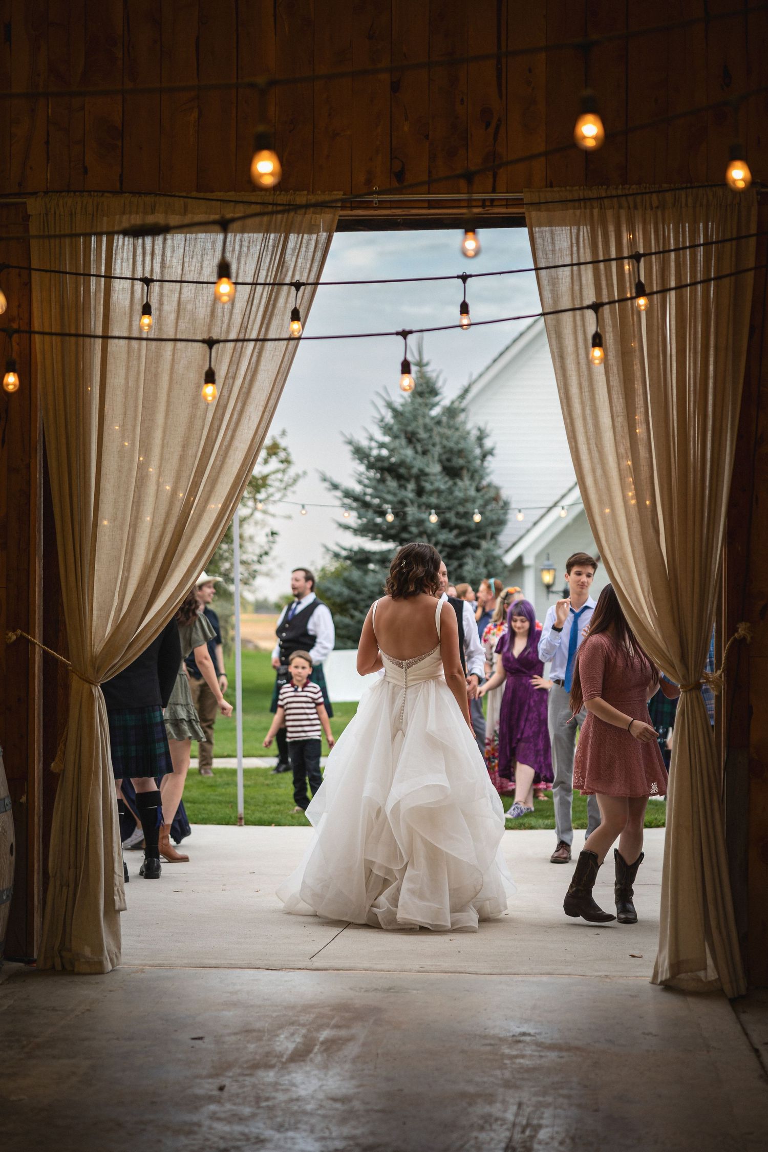 Wedding guests gather through curtained doorway at rustic venue with string lights overhead.