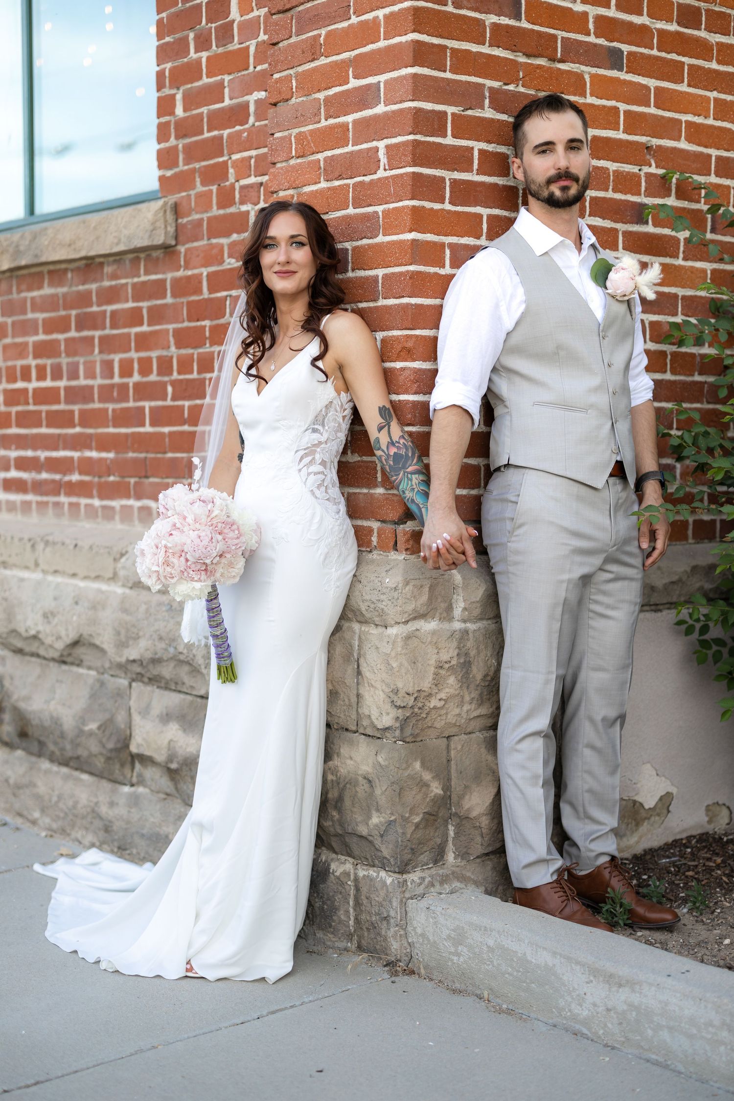 Modern wedding couple poses casually against stone and brick wall with pink bouquet.