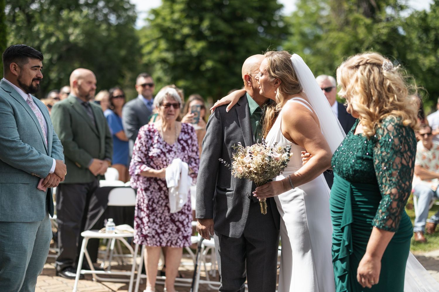 Wedding attendees gather outdoors at a summer ceremony under trees and blue skies.