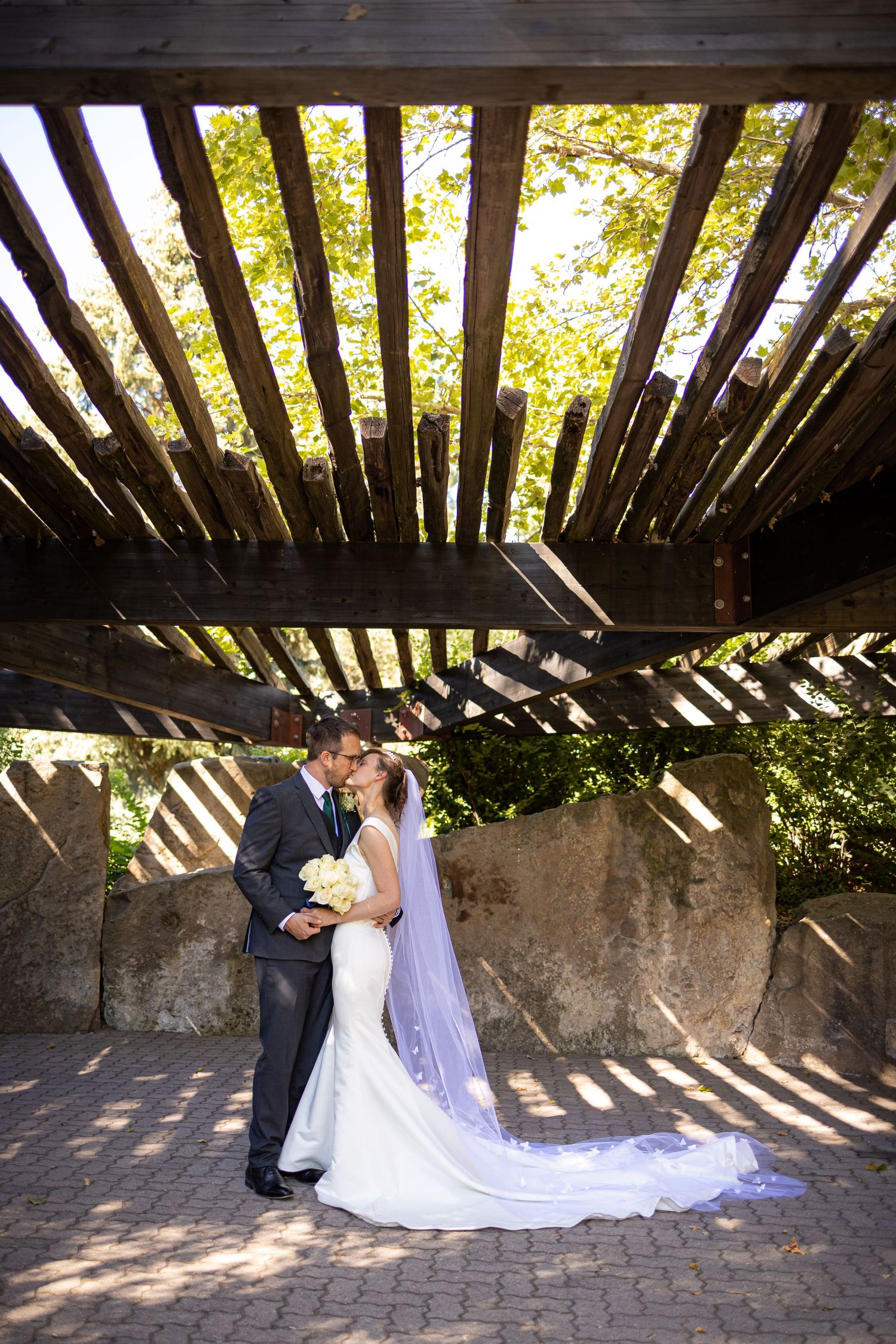 Newlyweds embrace under wooden pergola creating dramatic shadows on stone patio.