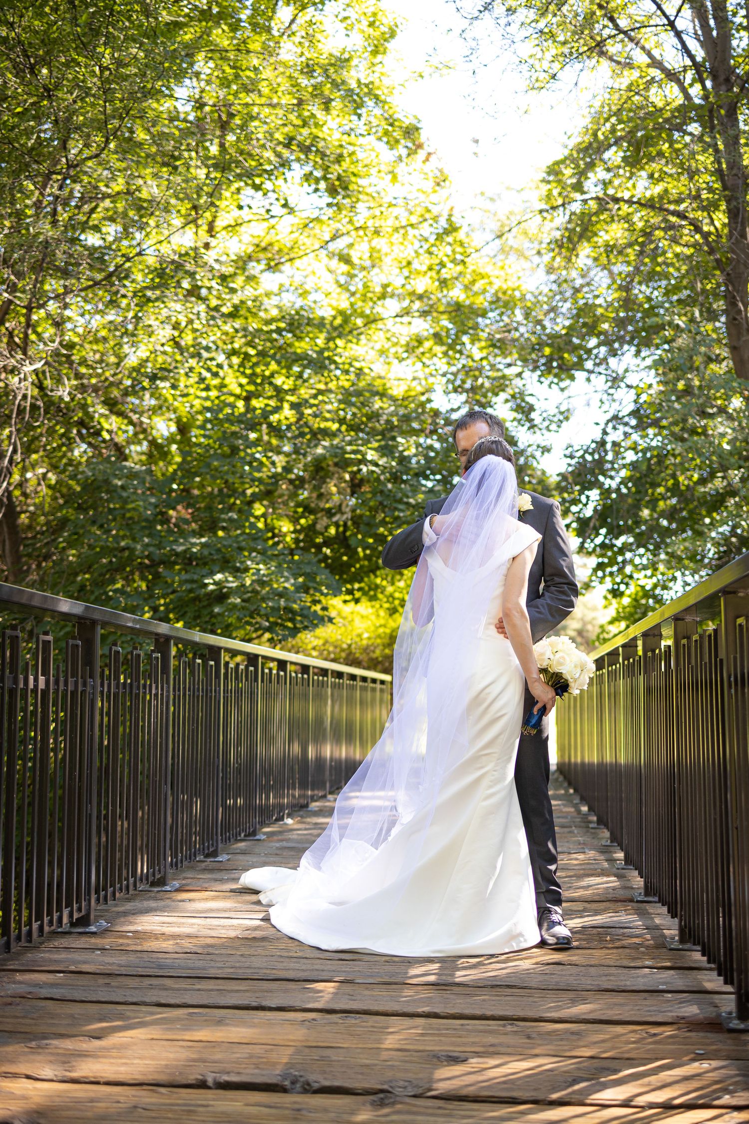 Romantic wedding portrait on wooden bridge with sunlight filtering through trees.