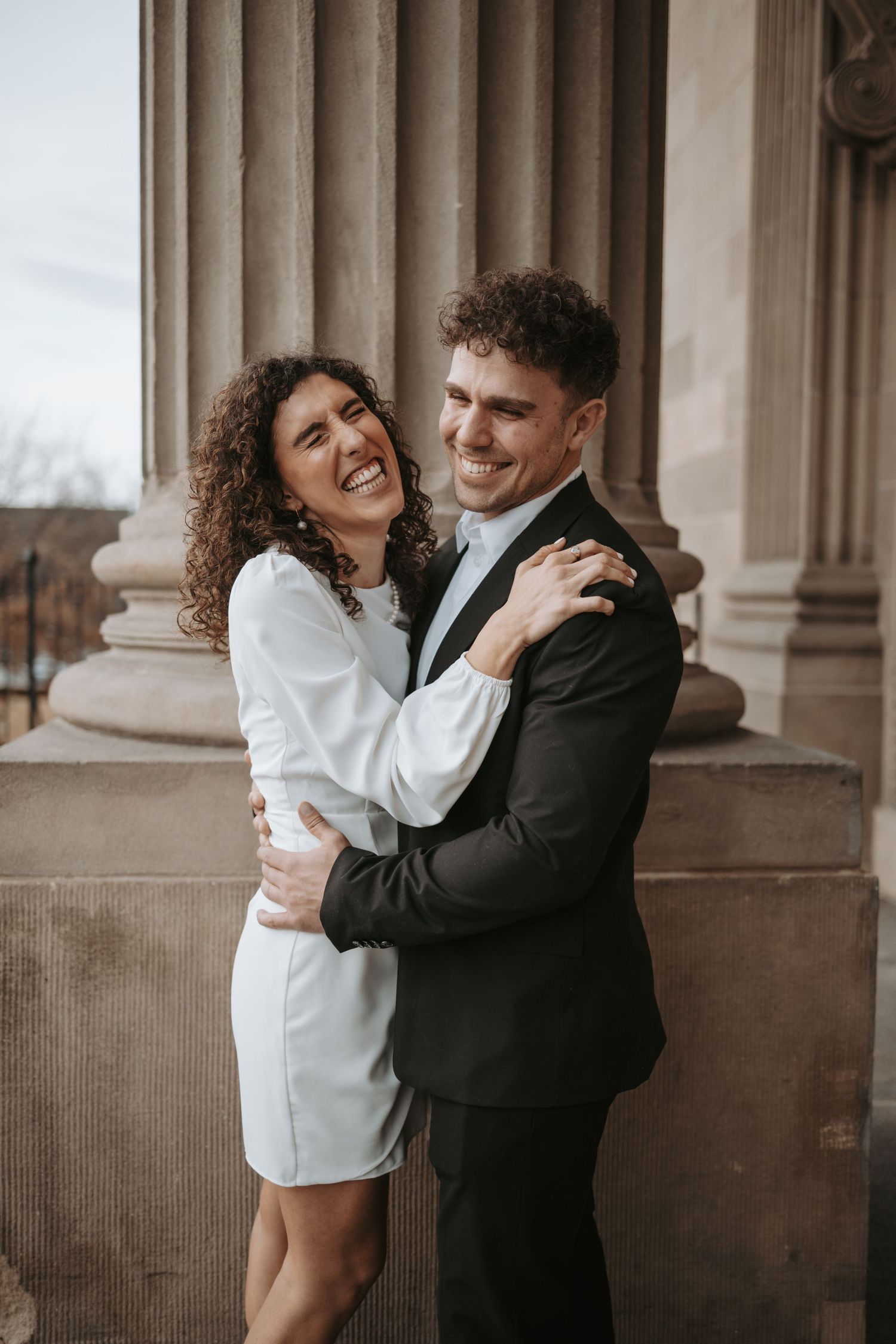 A couple embraces and laughs together while posing near stone columns in formal attire.