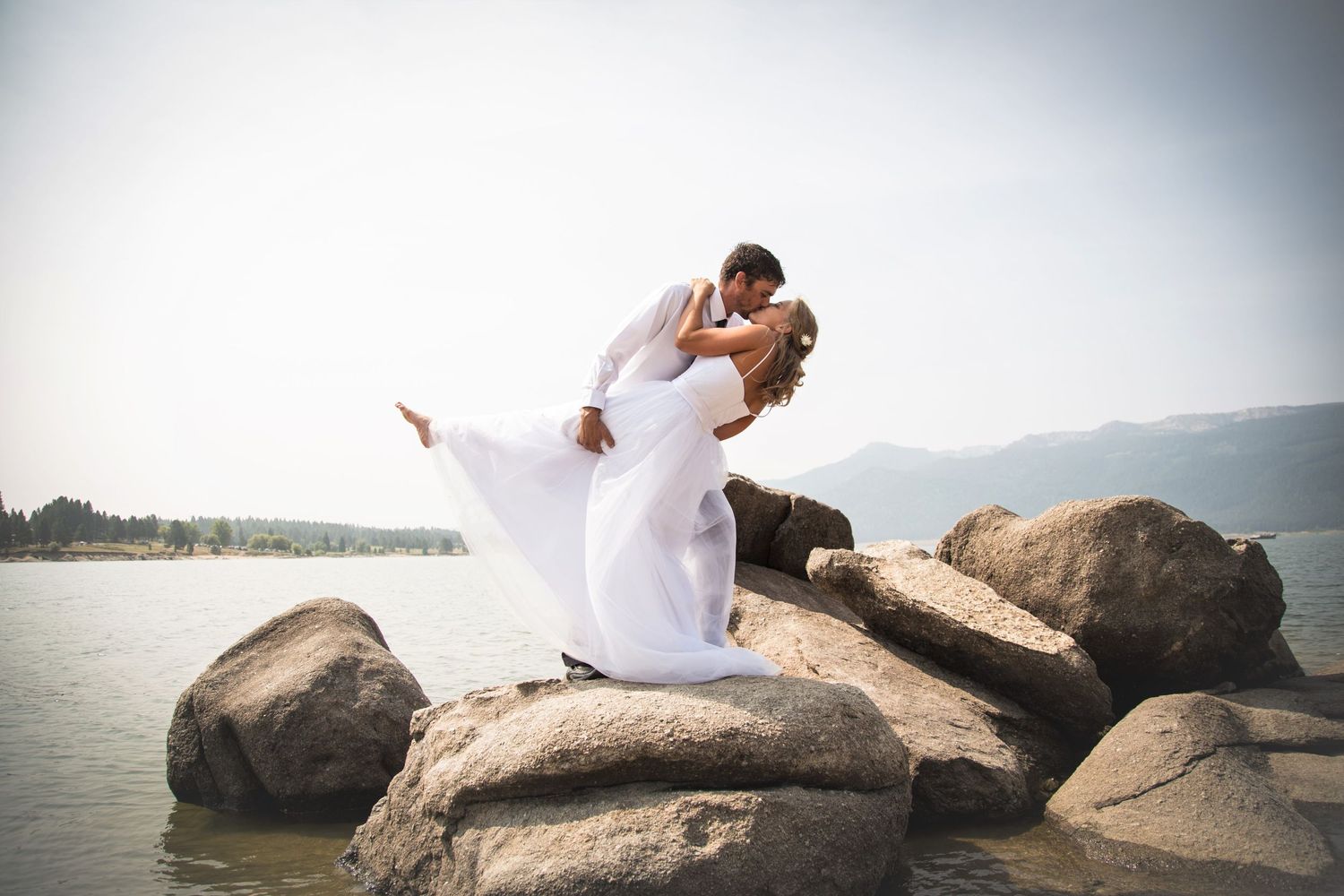 Dramatic wedding photo of couple embracing on rocky shoreline with lake backdrop.