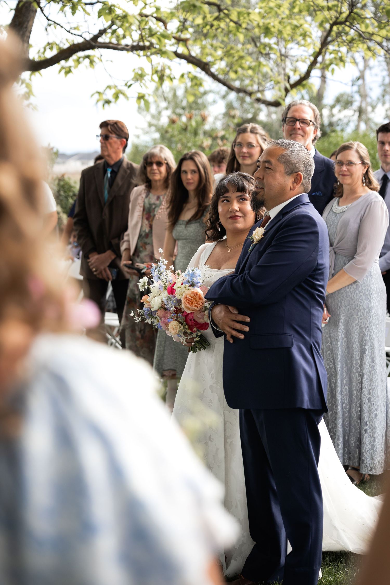 Wedding party and family members gather for outdoor ceremony under tree branches.