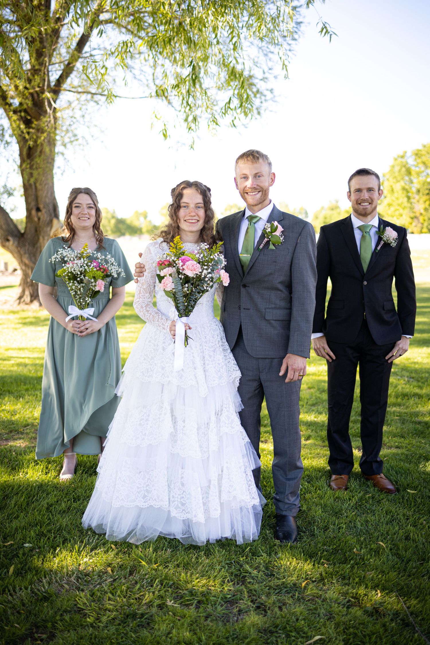 A wedding party poses on a sunny lawn with a bridesmaid in sage green and groomsman in dark suits with green ties.