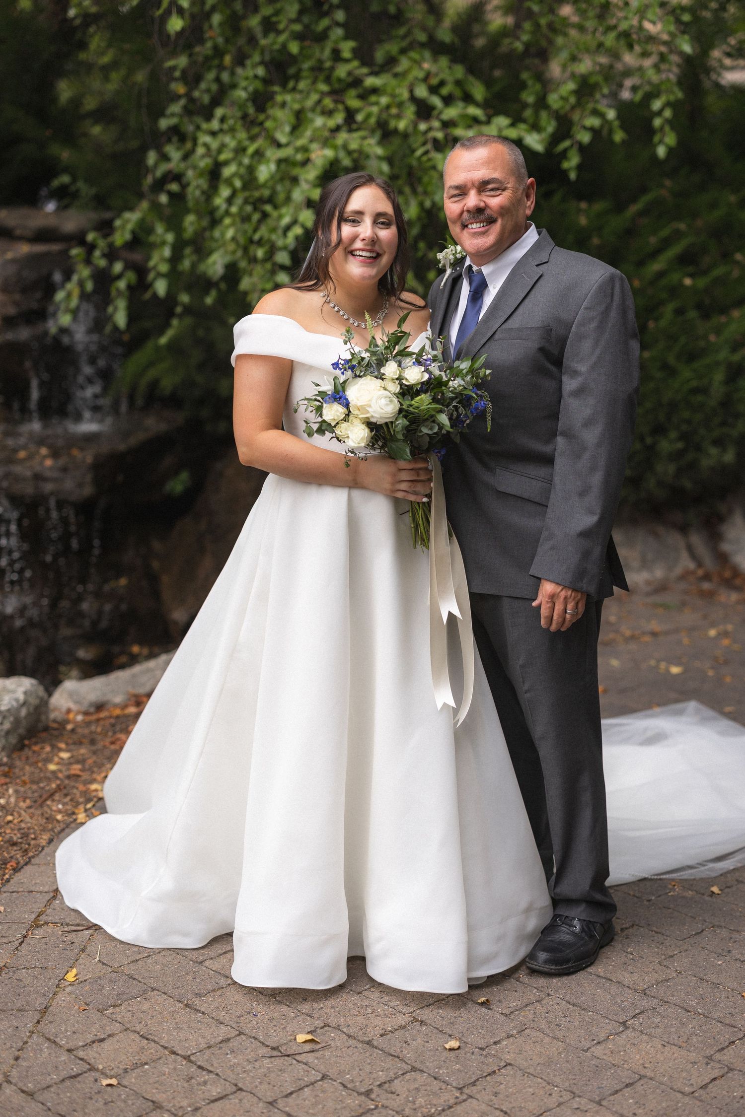 Happy wedding portrait in front of stone waterfall backdrop with white floral bouquet.