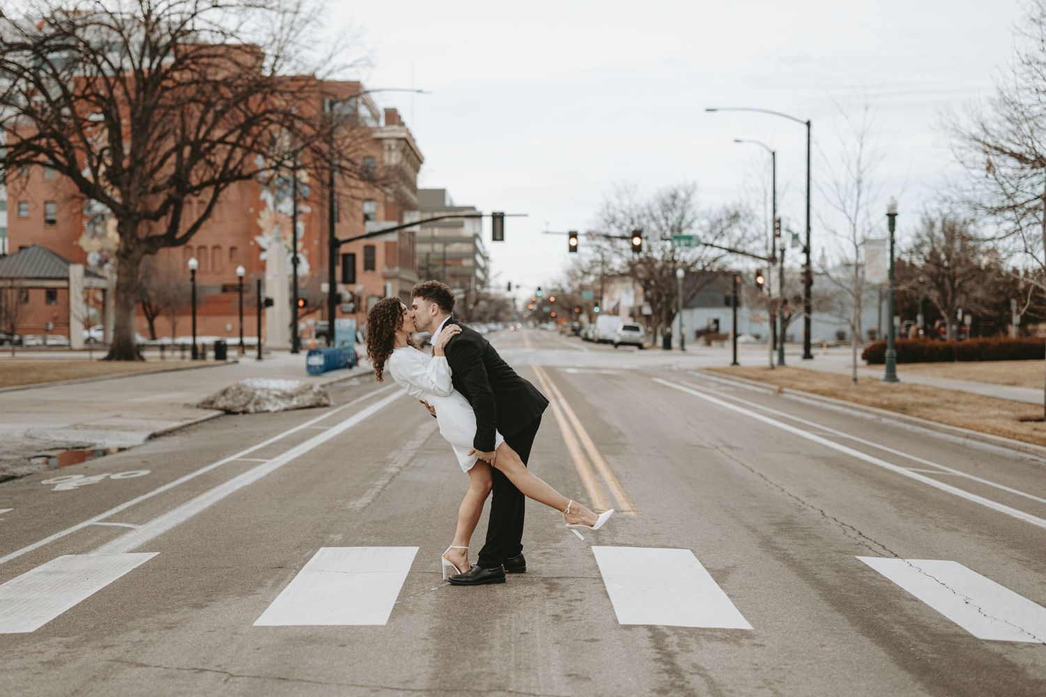 A couple share a romantic moment while crossing an empty city crosswalk in winter.