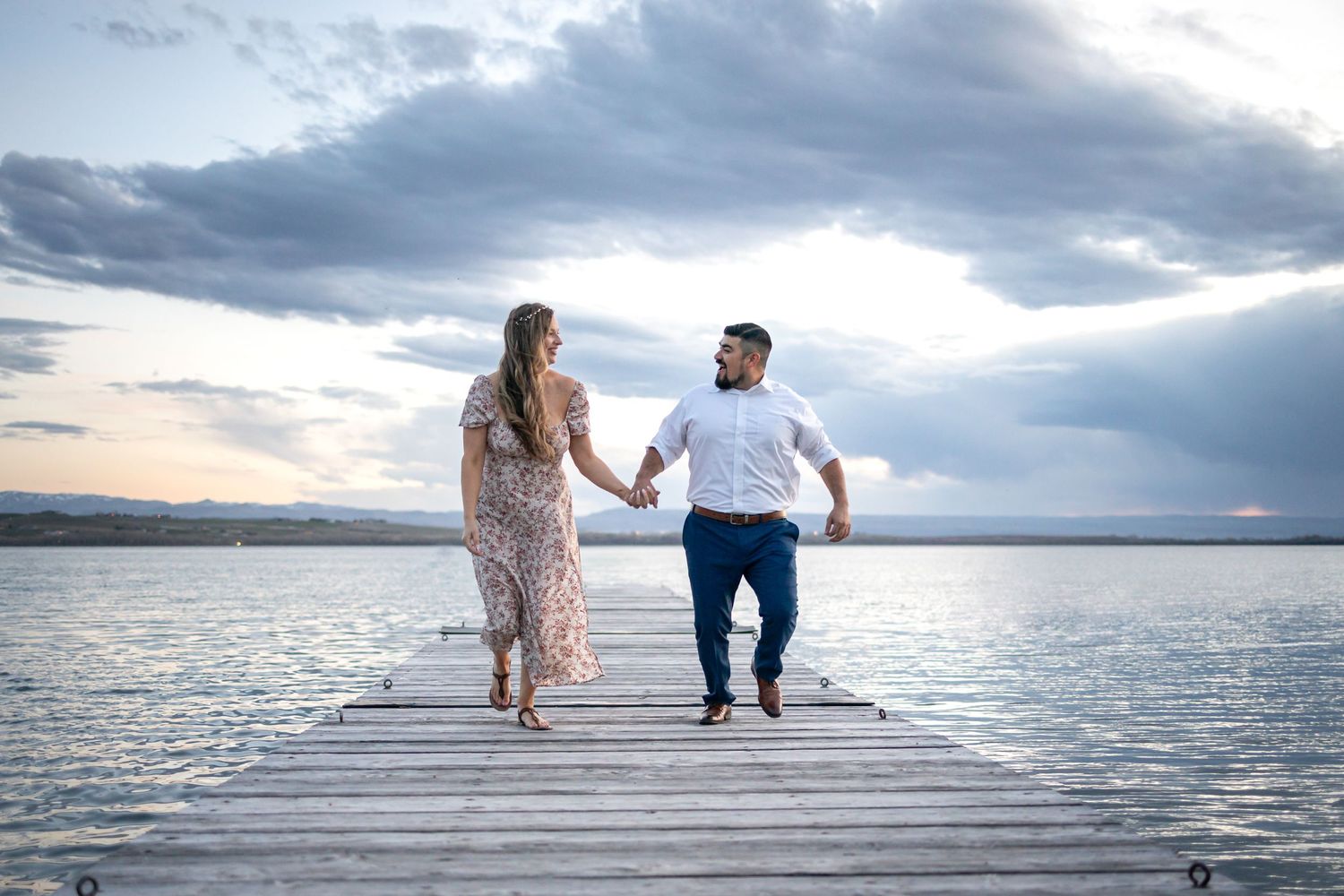 A couple holds hands while walking on a wooden dock over calm waters at sunset.
