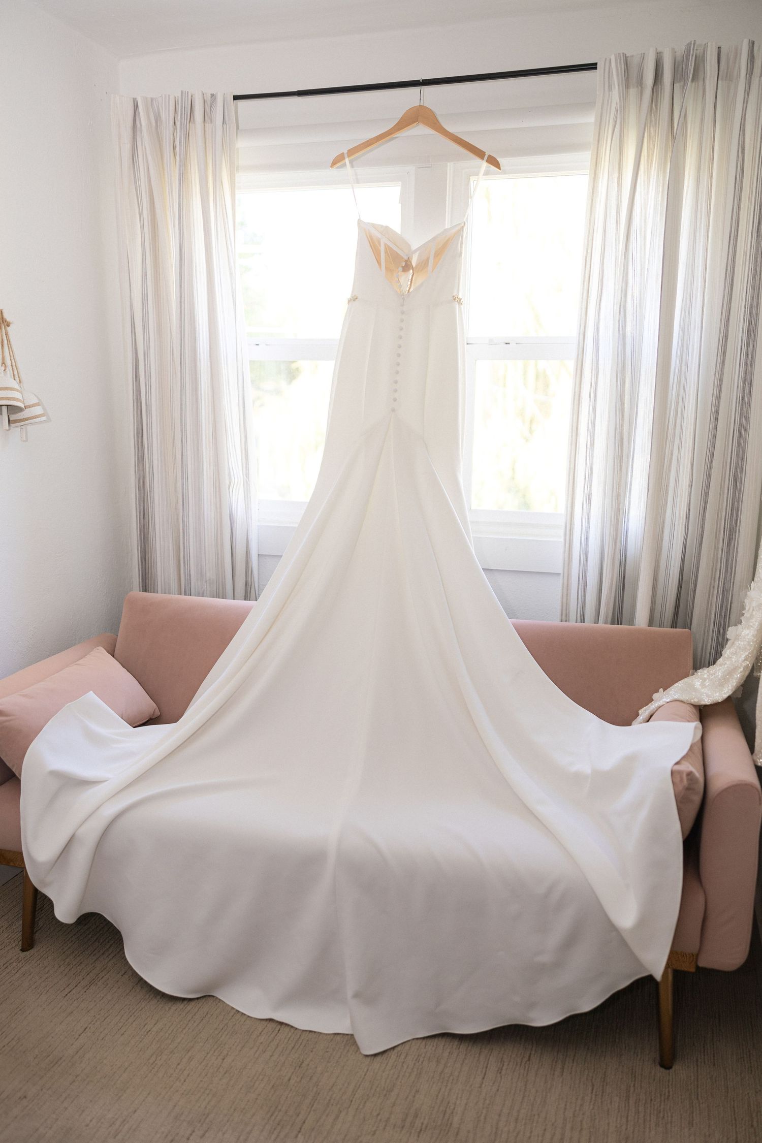 Flowing white wedding gown displayed on hanger in sunlit room with pink accent chair.