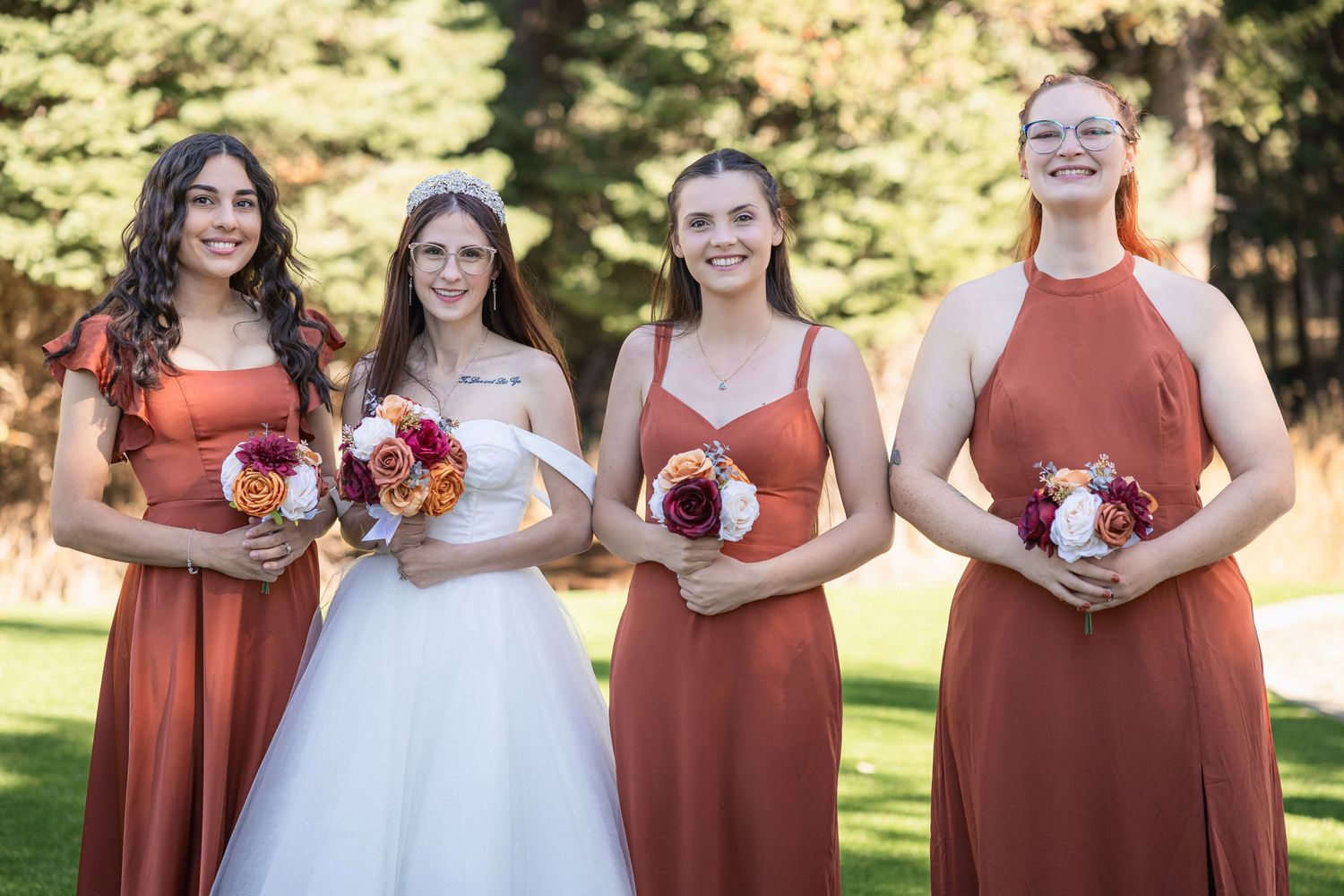 Wedding party poses outdoors with bridesmaids in terracotta dresses holding red bouquets.