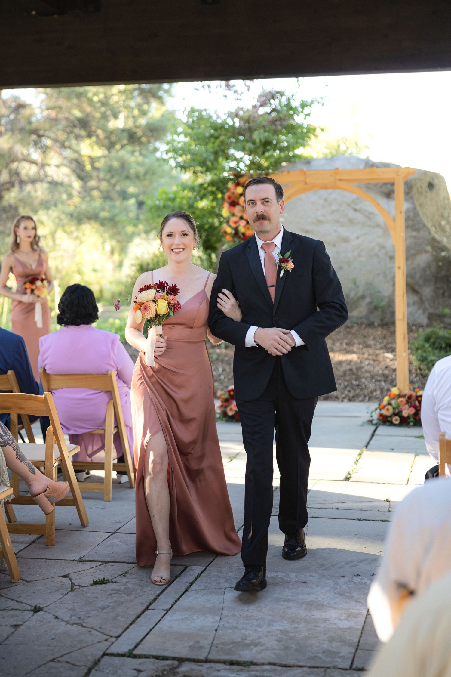 Wedding party member in flowing dusty rose dress walks with groomsman during ceremony procession.